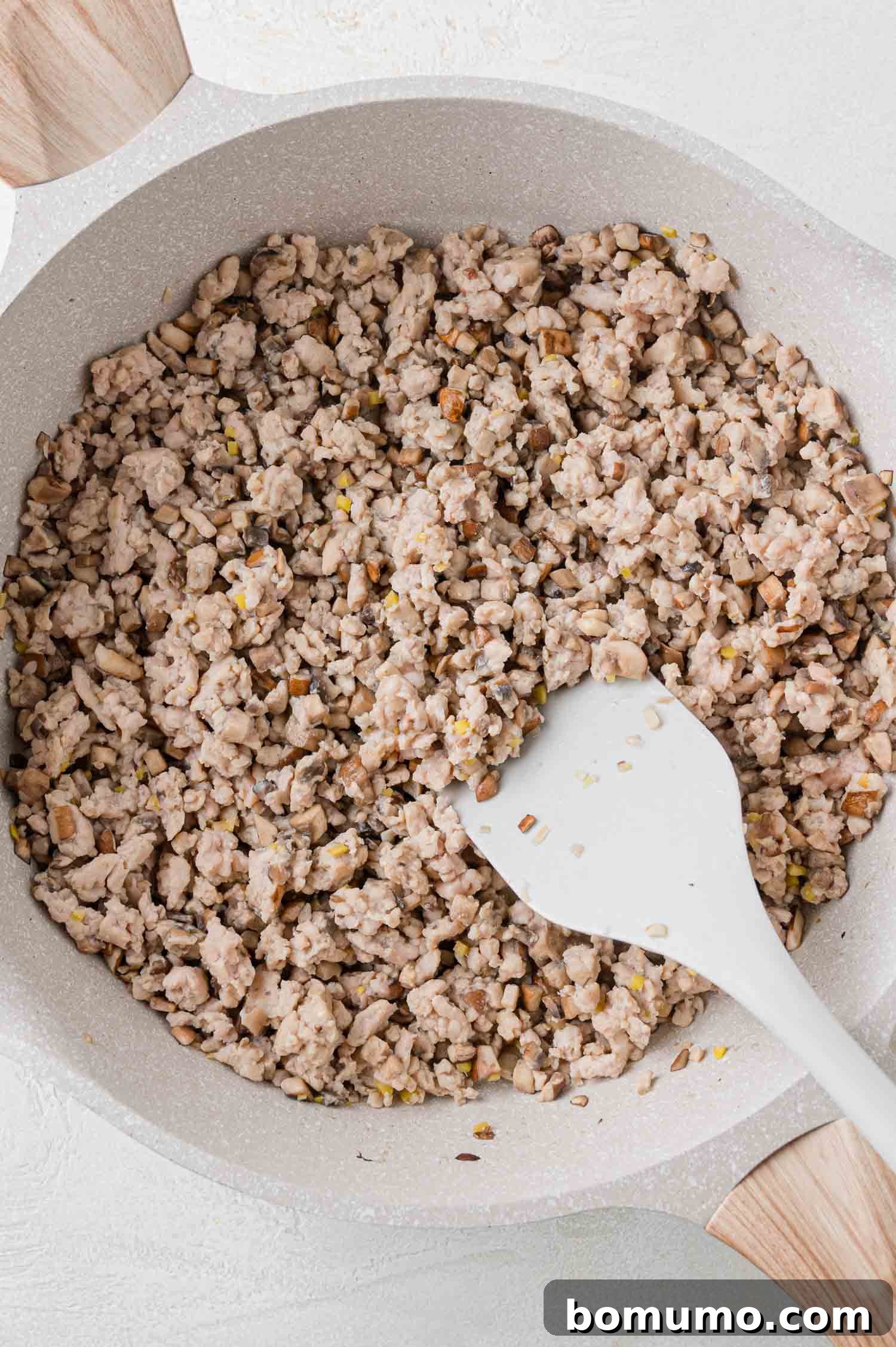 Pan filled with cooked ground meat and small bits of vegetables, stirred with a white spatula.
