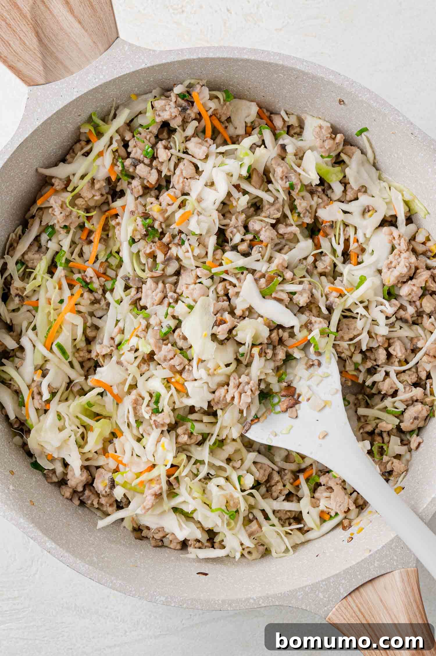 A skillet filled with a mixture of cooked ground meat, cabbage, carrots, and green onions, being stirred with a white spatula.
