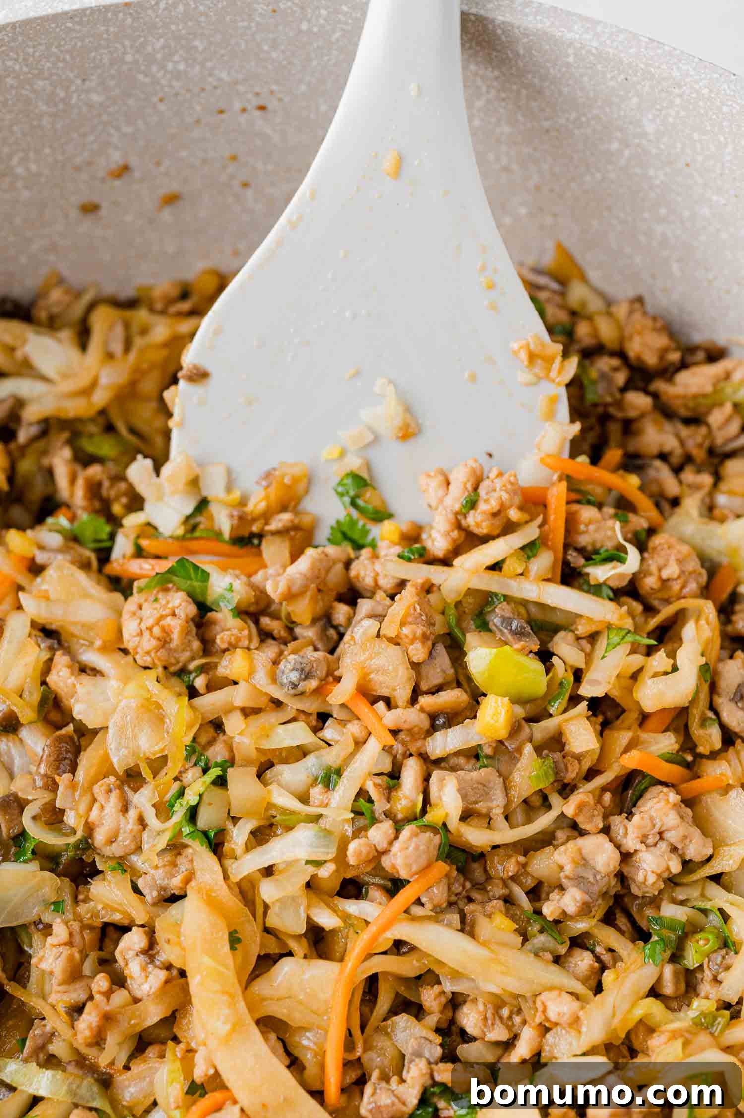 A close-up of a frying pan filled with chicken lettuce wrap filling containing ground chicken, shredded cabbage, carrots, and green herbs, being stirred with a white spatula.