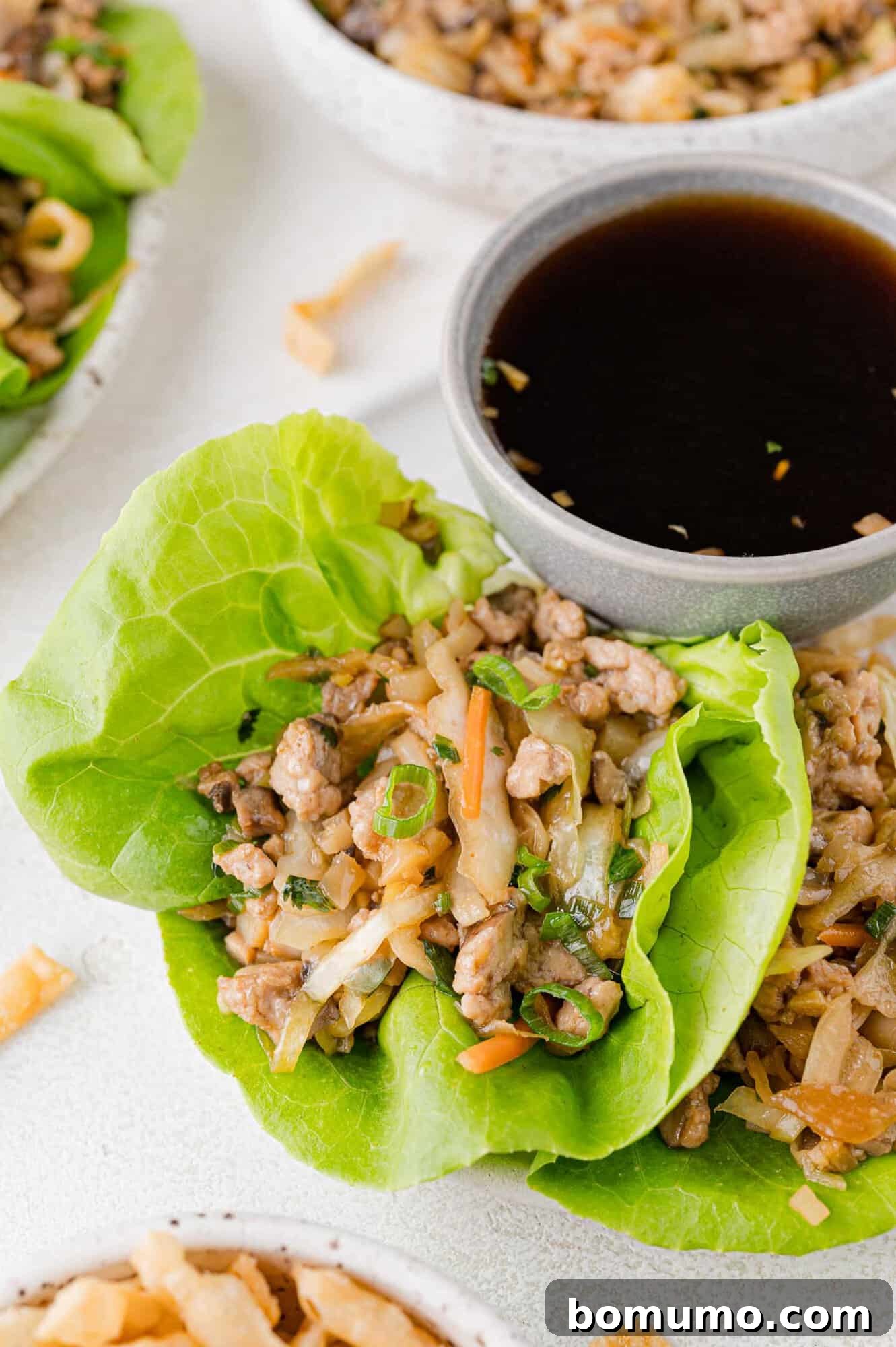 Lettuce wrap filled with ground meat and vegetables, served with a small bowl of dark dipping sauce on a white surface.