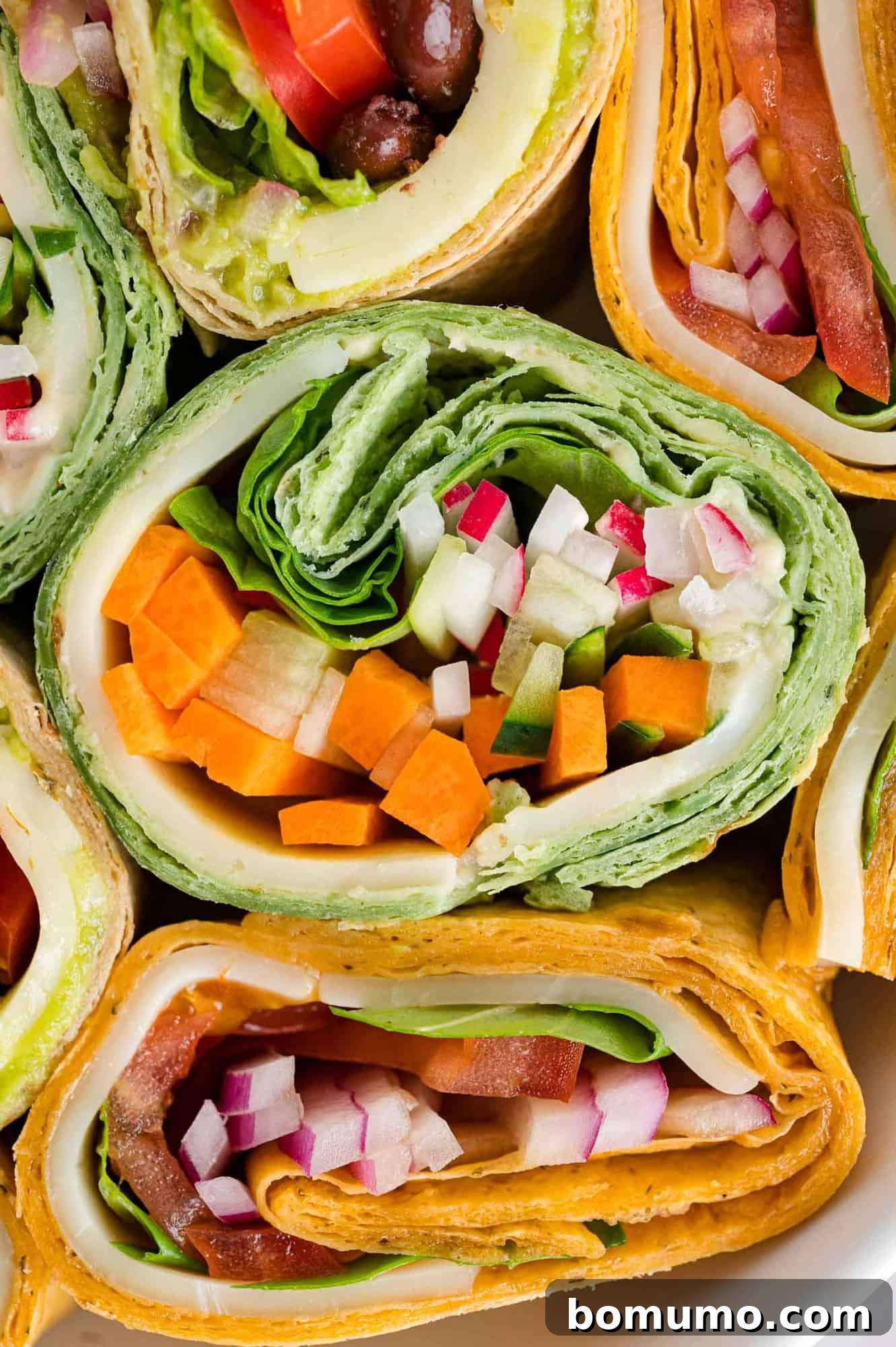 Cross section of multiple veggie wraps, showcasing colorful layers of vegetables, cheese, and hummus.