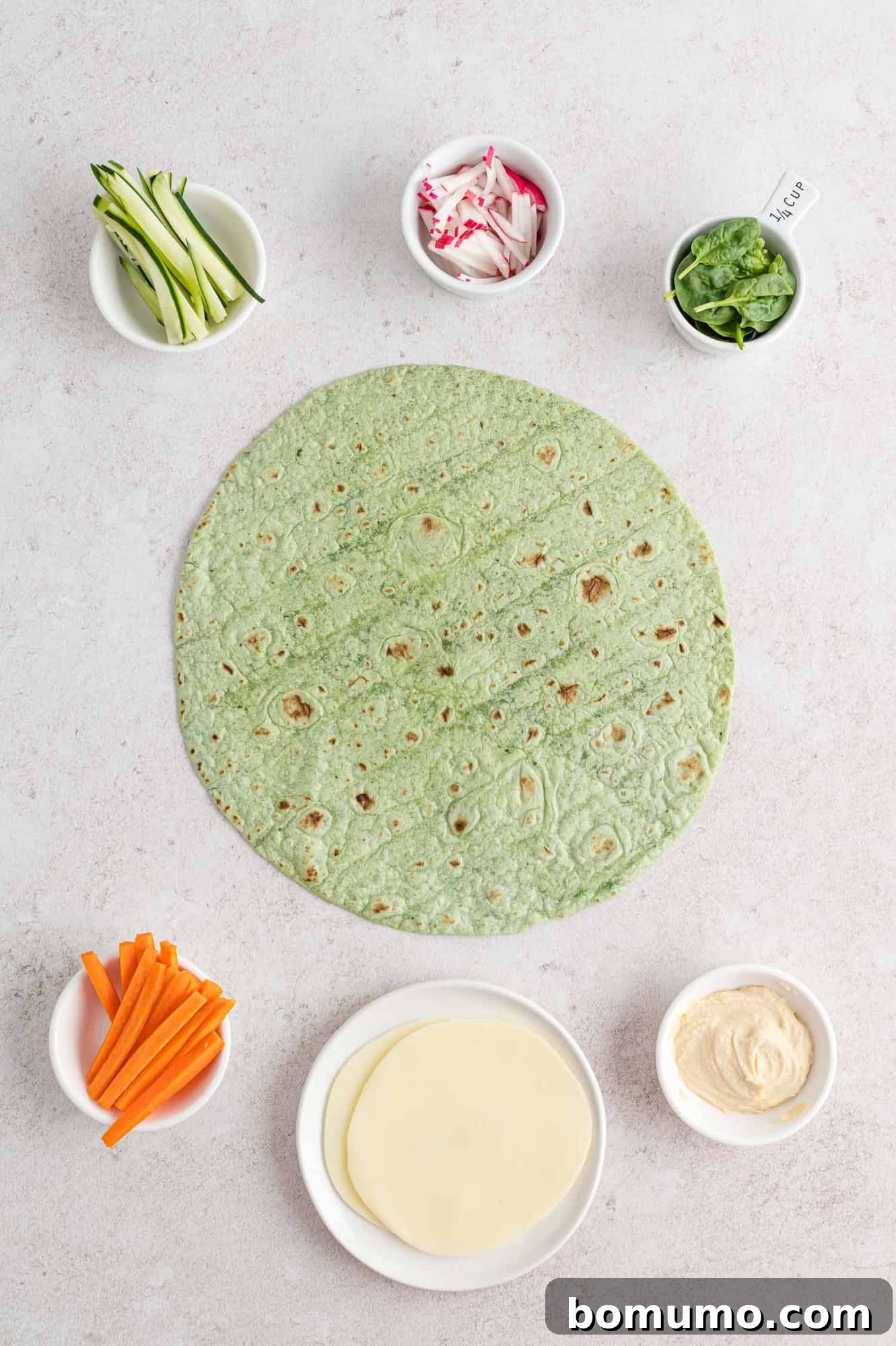 Assorted fresh ingredients for making a veggie wrap laid out on a cutting board, including tortillas, hummus, cheese, and various vegetables.