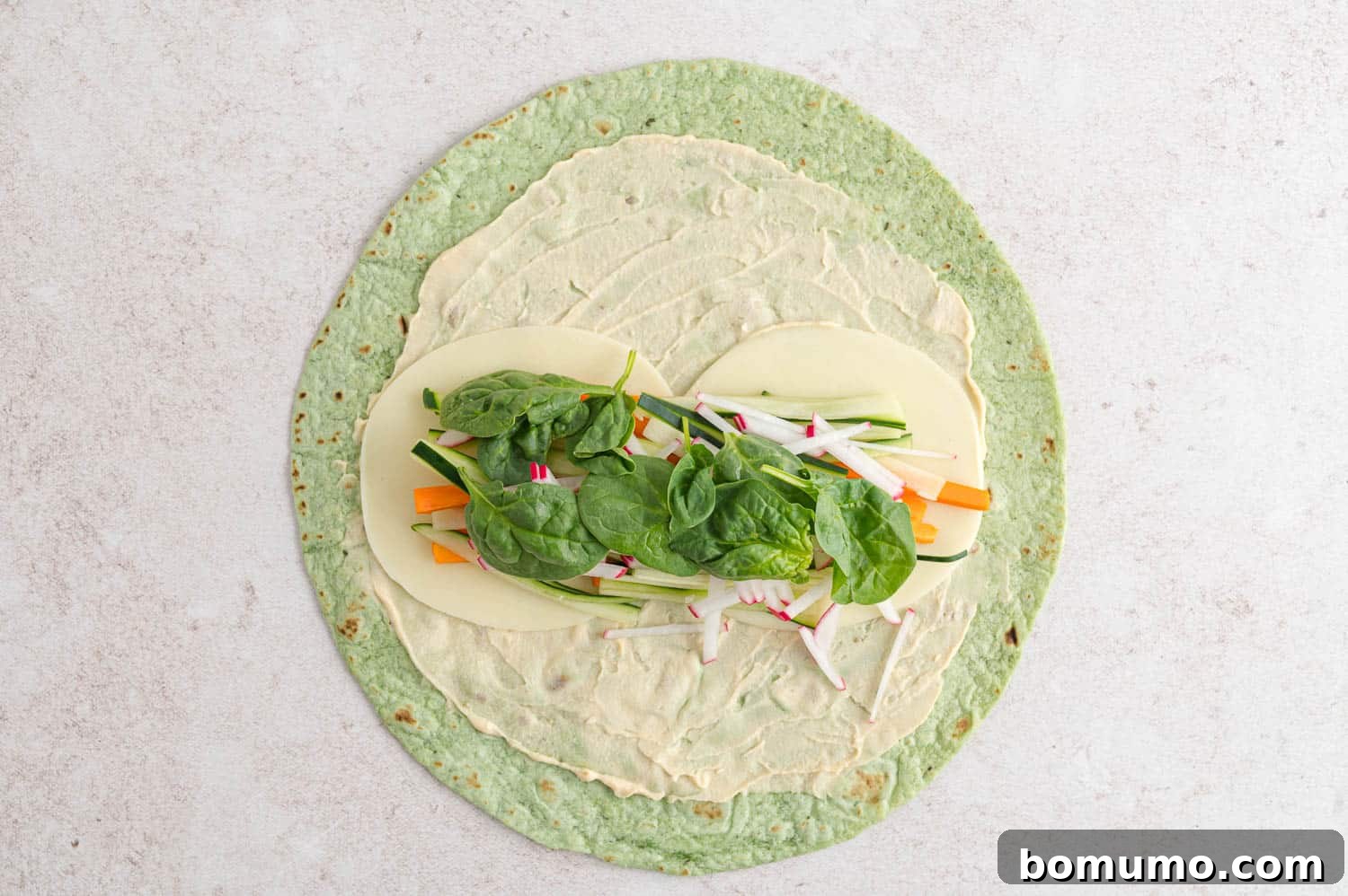 An unrolled tortilla with hummus, sliced cheese, julienned carrots, cucumbers, radishes, and spinach layered on one side.
