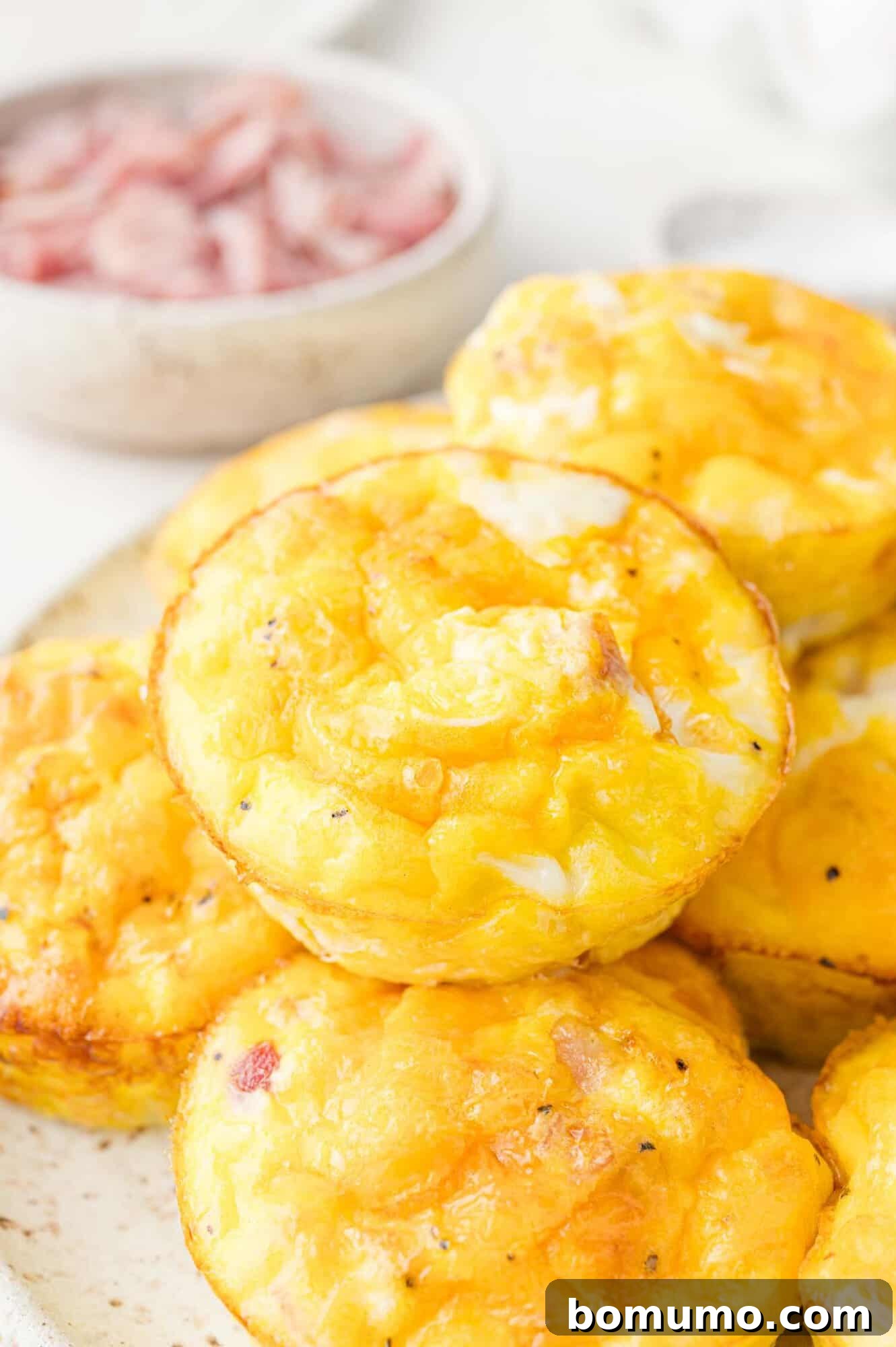 A plate of baked egg muffins with visible melted cheese and diced pieces of ham. A small bowl of additional diced ham is in the background.