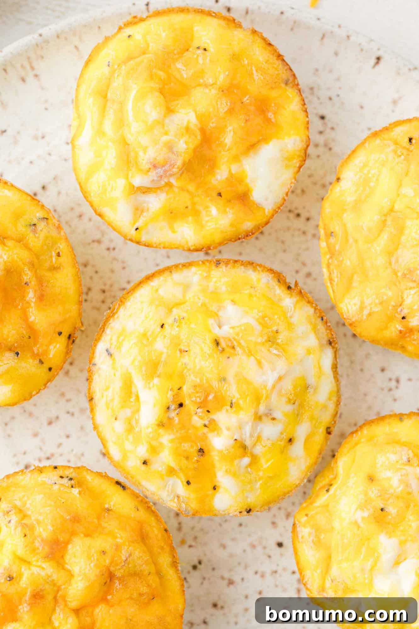 Close-up of six golden brown egg muffins with visible cheese and seasoning on a speckled plate.