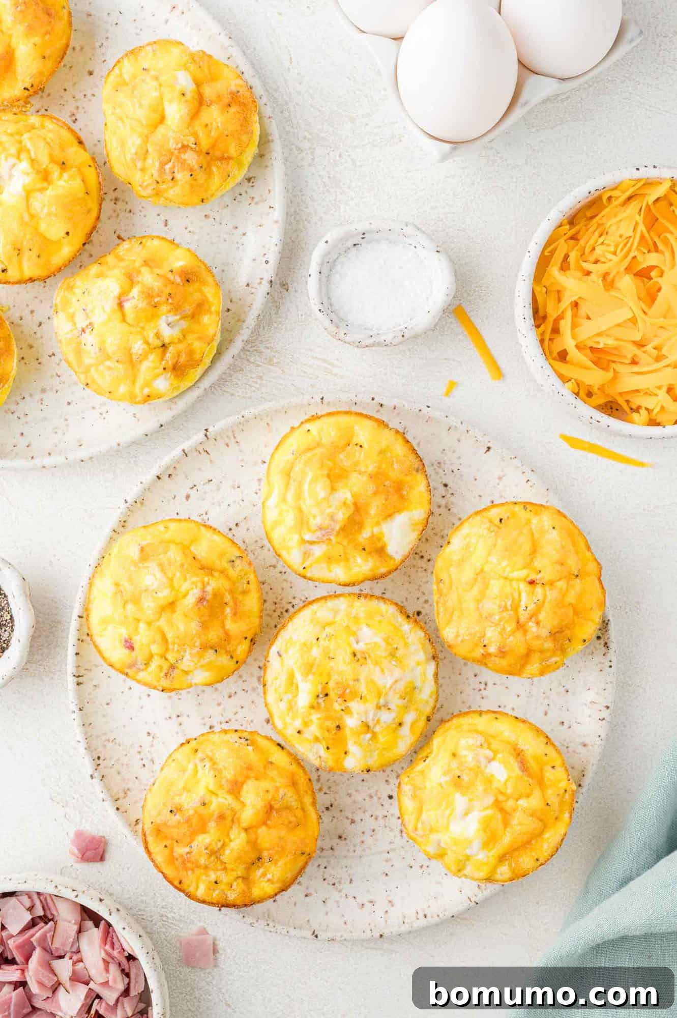 Plates of baked egg muffins with cheese on a light surface, surrounded by a bowl of shredded cheese, salt, whole eggs, and chopped ham.