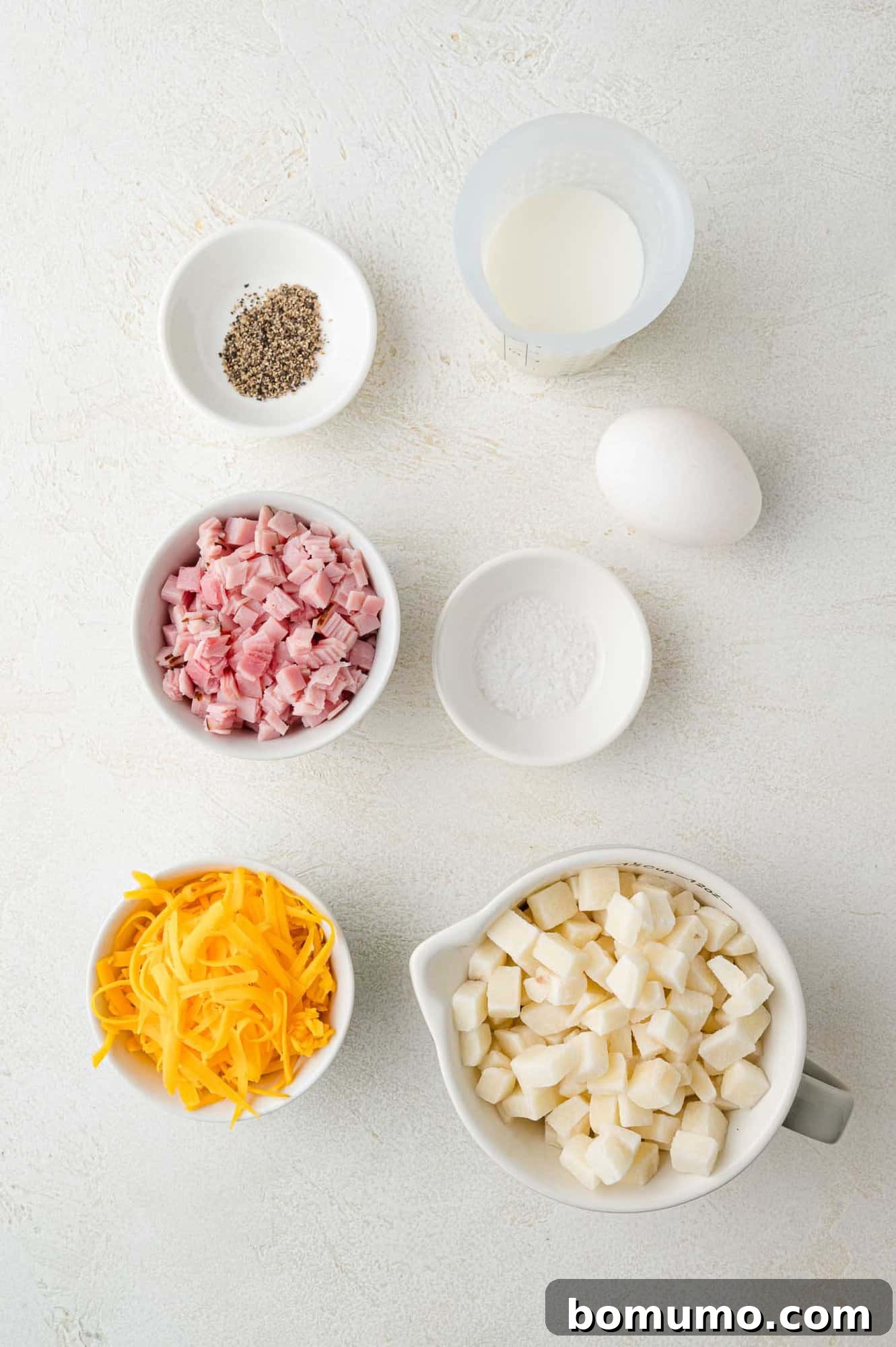 Ingredients arranged on a white surface: diced ham, shredded cheese, diced potatoes, a whole egg, milk, pepper, and salt.