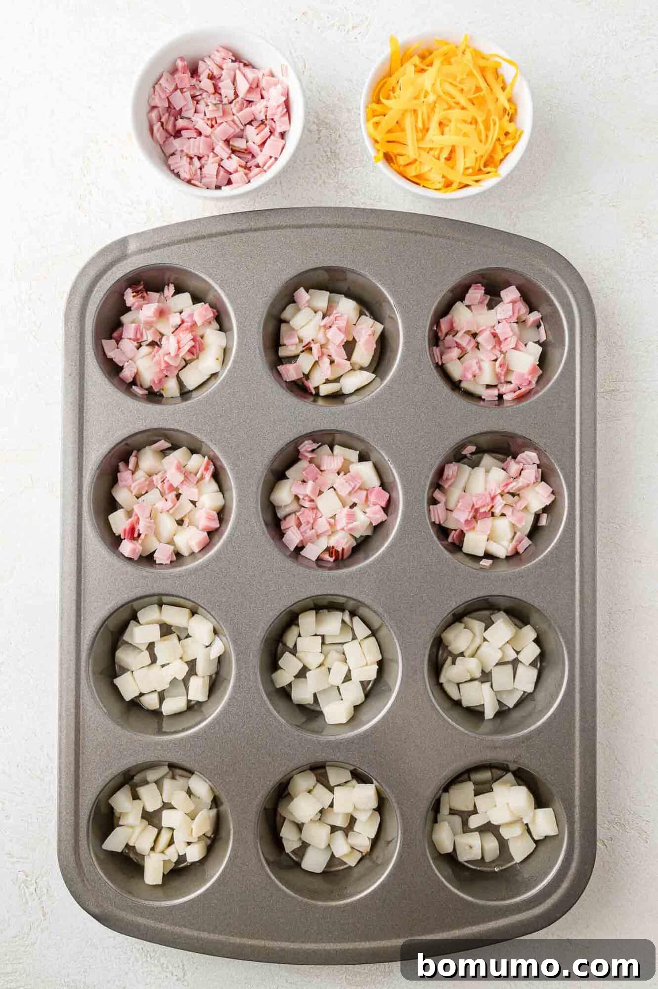 A muffin tin with diced ham and potatoes in each cup. Nearby are bowls of extra diced ham and shredded cheddar cheese.