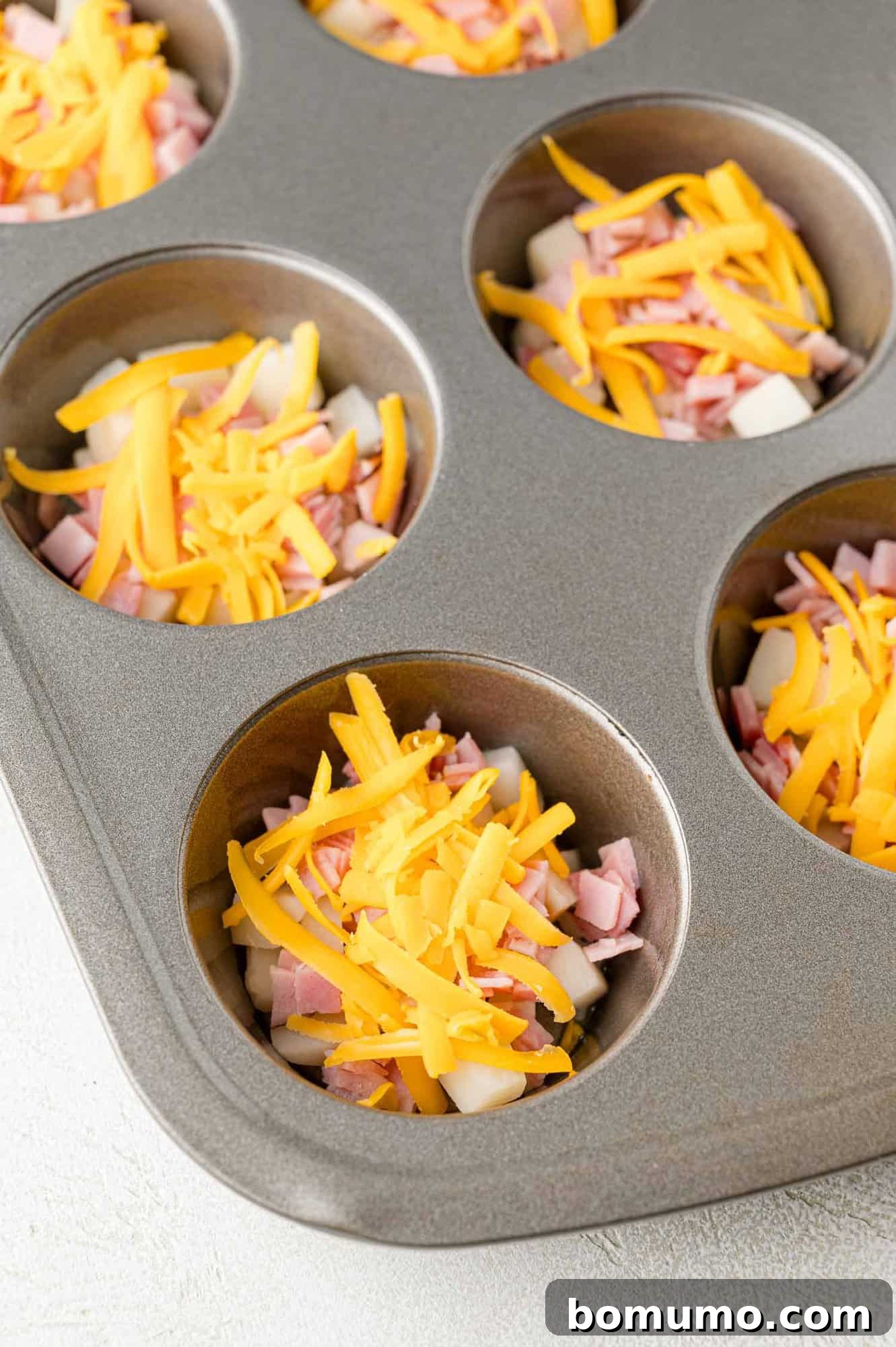 A muffin tin filled with diced ham, potatoes, and shredded cheese in each compartment.