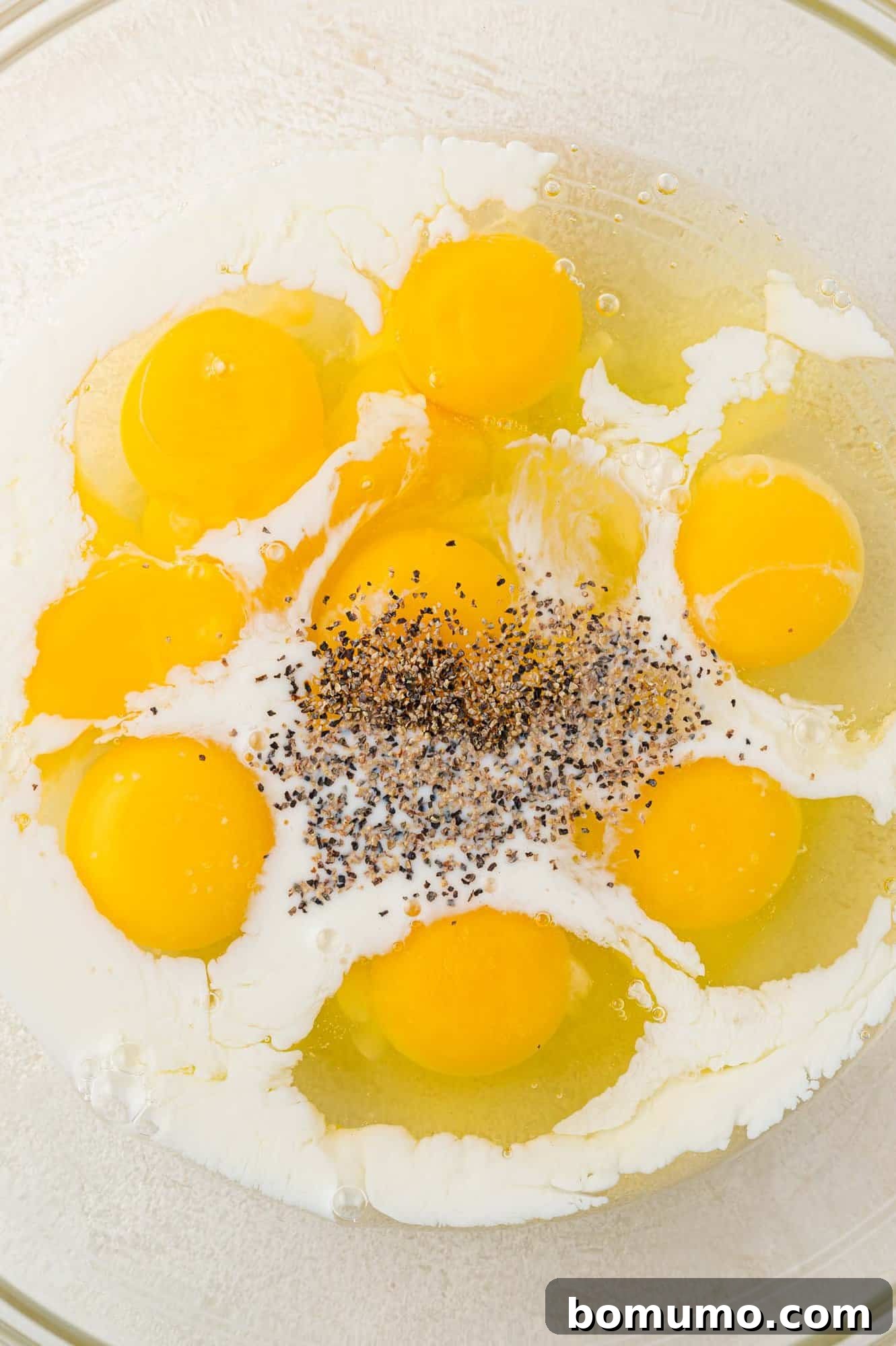 A glass bowl contains raw eggs, milk, and ground pepper.