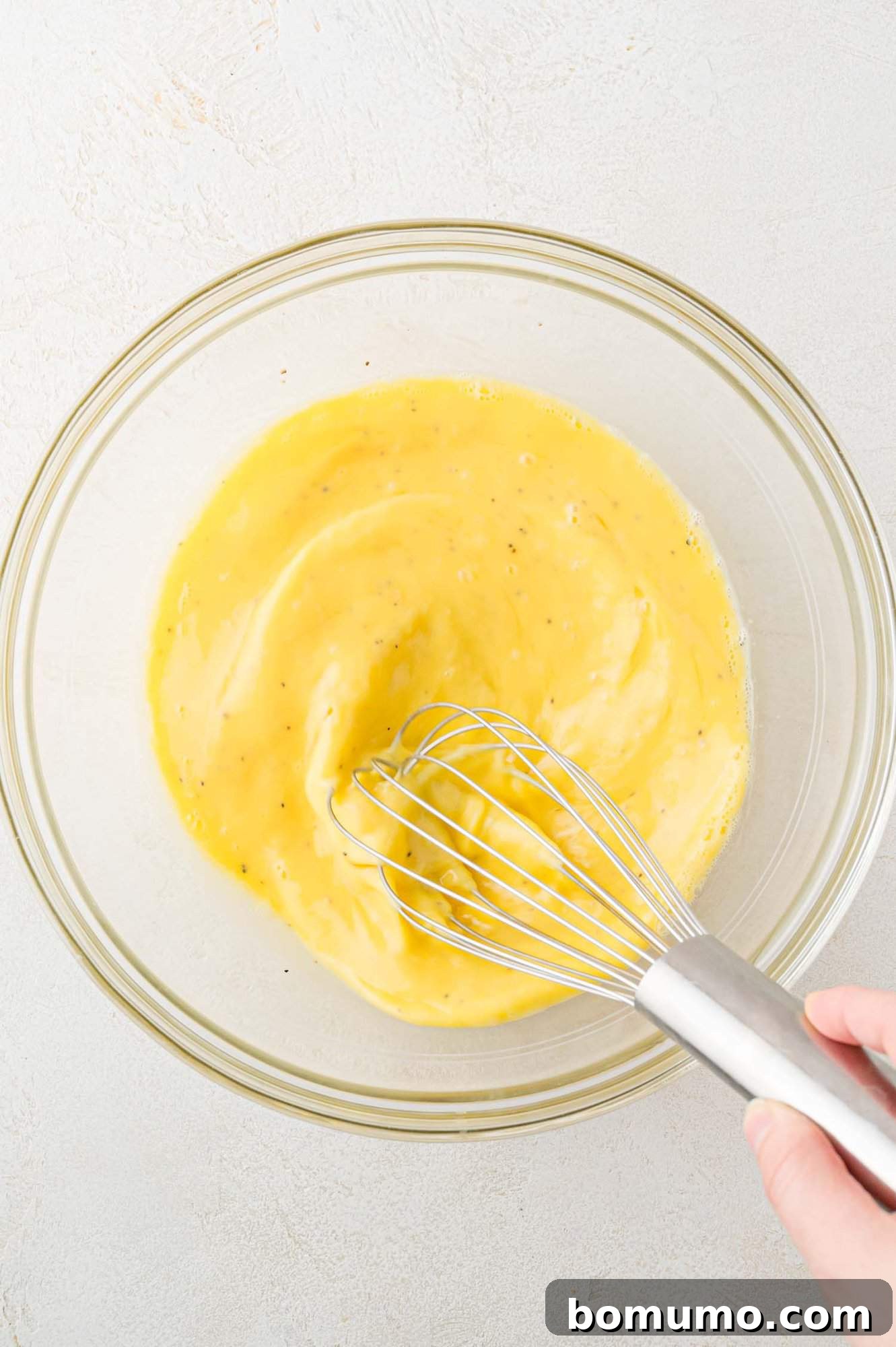 A hand is whisking beaten eggs in a clear glass bowl on a light gray surface.
