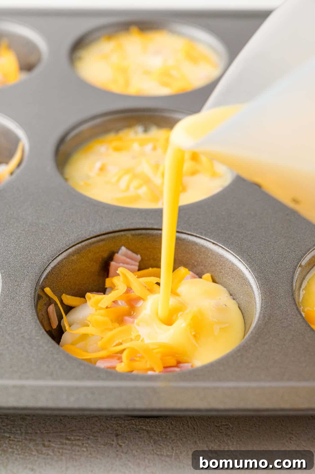 Egg mixture being poured into muffin tin with shredded cheese and meat.
