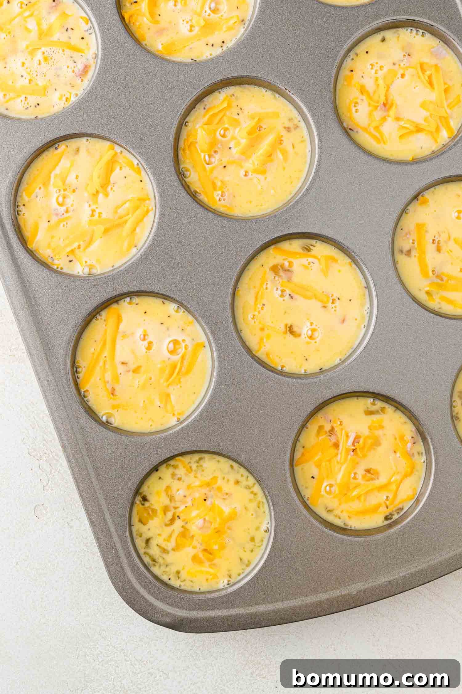 A muffin tin filled with uncooked egg mixture, and shredded cheese, ready to be baked.
