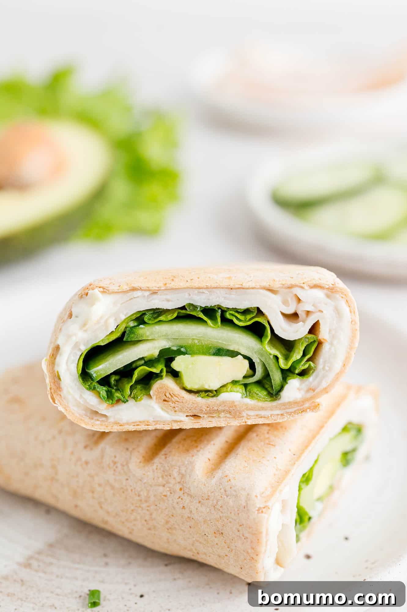 Two halves of a vibrant turkey wrap presented on a white plate, with a fresh avocado and a bowl of crisp cucumber slices in the background. A perfect illustration of a healthy and appealing meal.