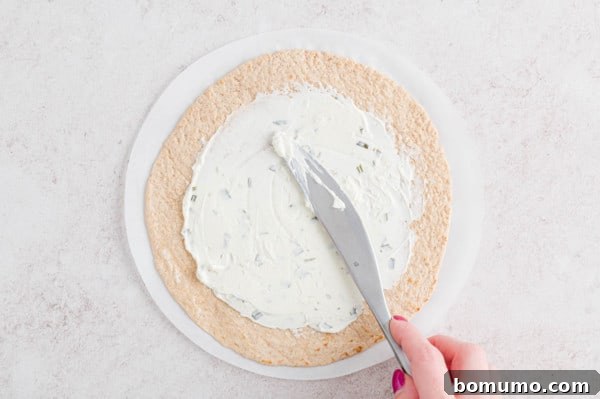 A skilled hand expertly spreading savory chive and onion cream cheese across a soft flour tortilla, meticulously preparing the essential base for a truly delicious and satisfying wrap.