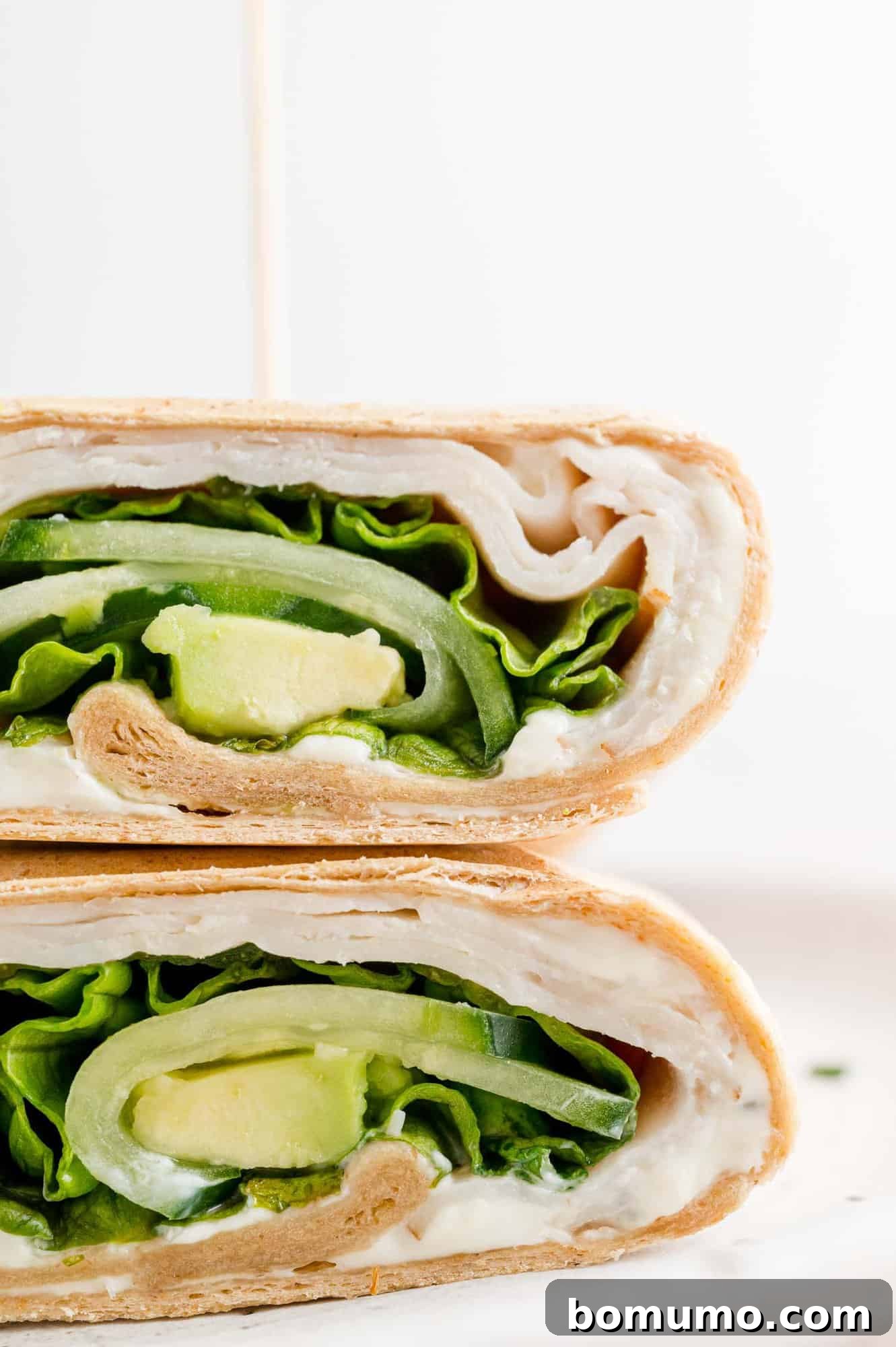 A close-up photograph showcasing two perfectly sliced halves of a turkey wrap, elegantly stacked on a pristine white plate, with all the delicious and colorful layers beautifully revealed.