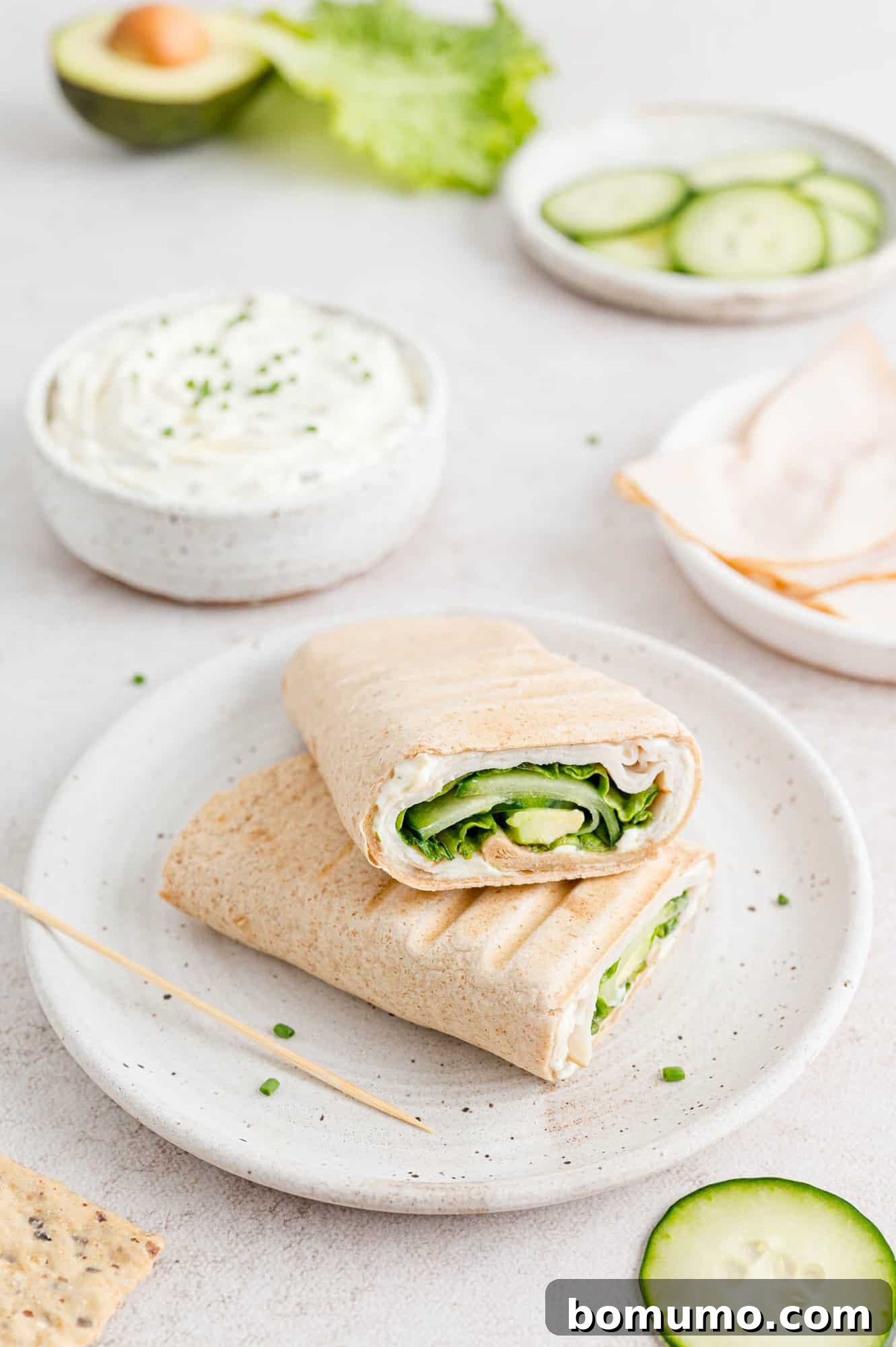 Two neatly presented halves of a turkey wrap on a white plate, artfully surrounded by small bowls filled with fresh, vibrant ingredients, illustrating the myriad customization options available.