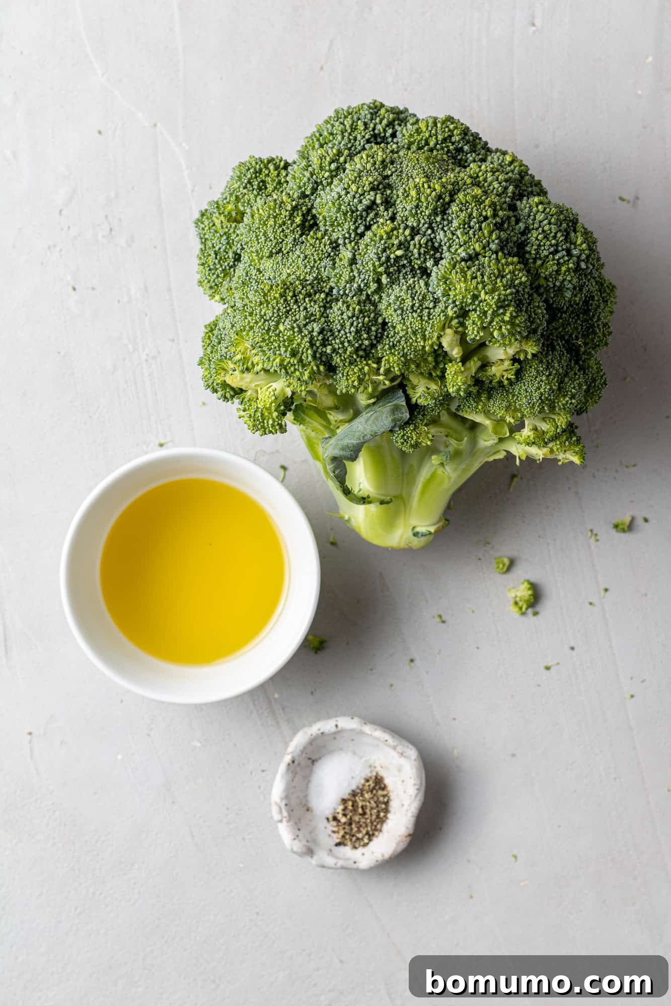 Overhead view of a fresh head of broccoli, alongside bottles of olive oil, and dishes of salt and pepper – all the simple ingredients needed for this recipe.