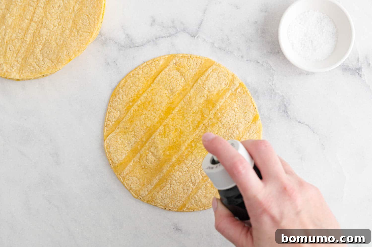 A hand holds an olive oil spray bottle, lightly misting a stack of corn tortillas on a cutting board before they are cut into chips.