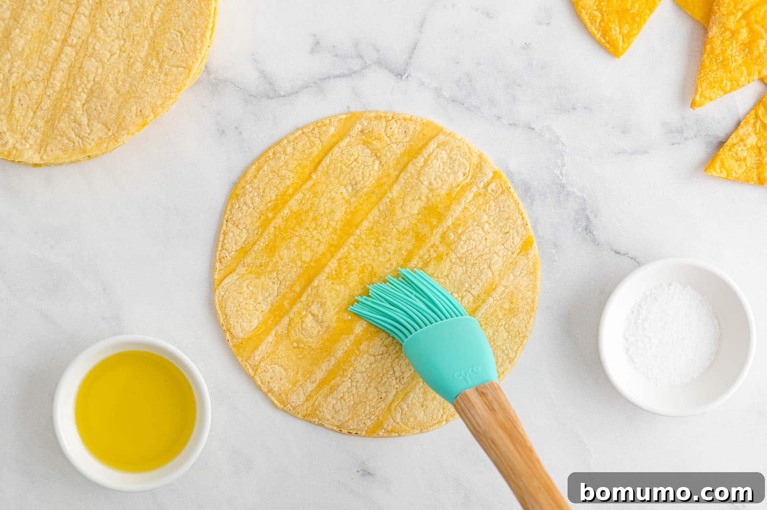 A pastry brush applies a thin layer of olive oil to a round corn tortilla, showcasing an alternative method to oil spray.