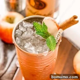 An iced drink in a copper cup, garnished with apple, mint leaves, and a cinnamon stick