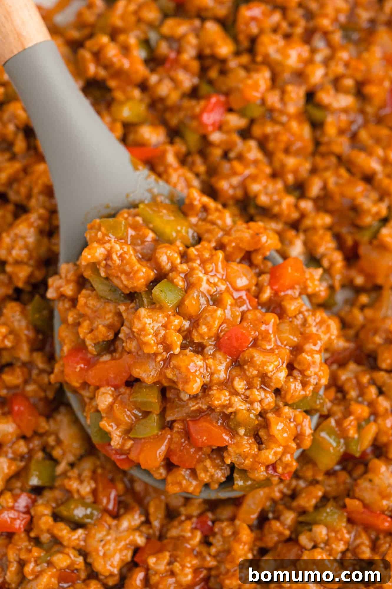 A large serving spoon scooping up chunky, rich sloppy Joe meat, highlighting the vibrant mixture of ground meat, bell peppers, and onions in a savory sauce.