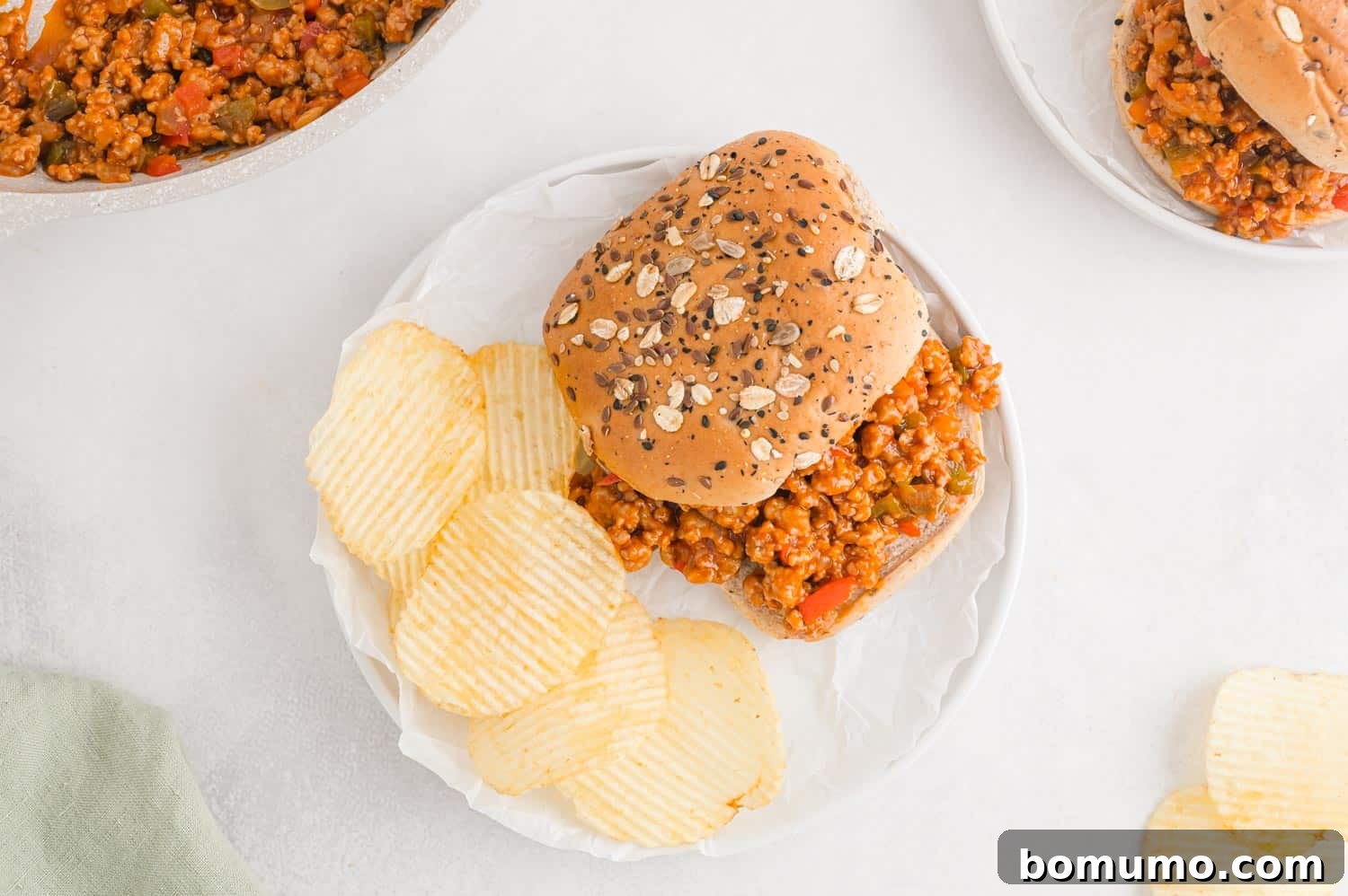 A delectable sloppy Joe sandwich served on a white plate, accompanied by a bowl of crispy potato chips, creating a classic and satisfying meal.