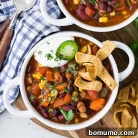 An overhead view of vegetarian chili in a white bowl, garnished with Fritos, sour cream, and jalapeños.