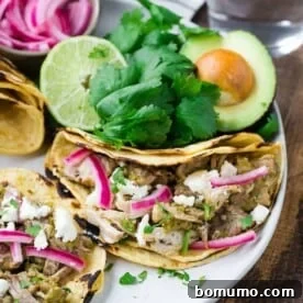 Three delicious pork tacos on a plate, garnished with fresh cilantro, avocado slices, and pickled red onions.