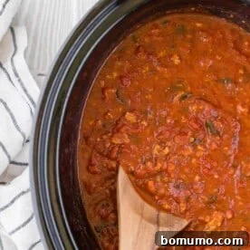A wooden spoon rests in a black crockpot filled with rich, simmering spaghetti sauce.