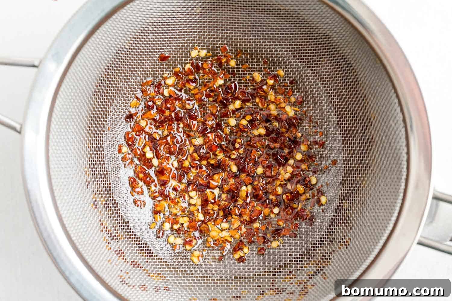 Red pepper flakes being strained from the hot honey mixture using a fine-mesh strainer.