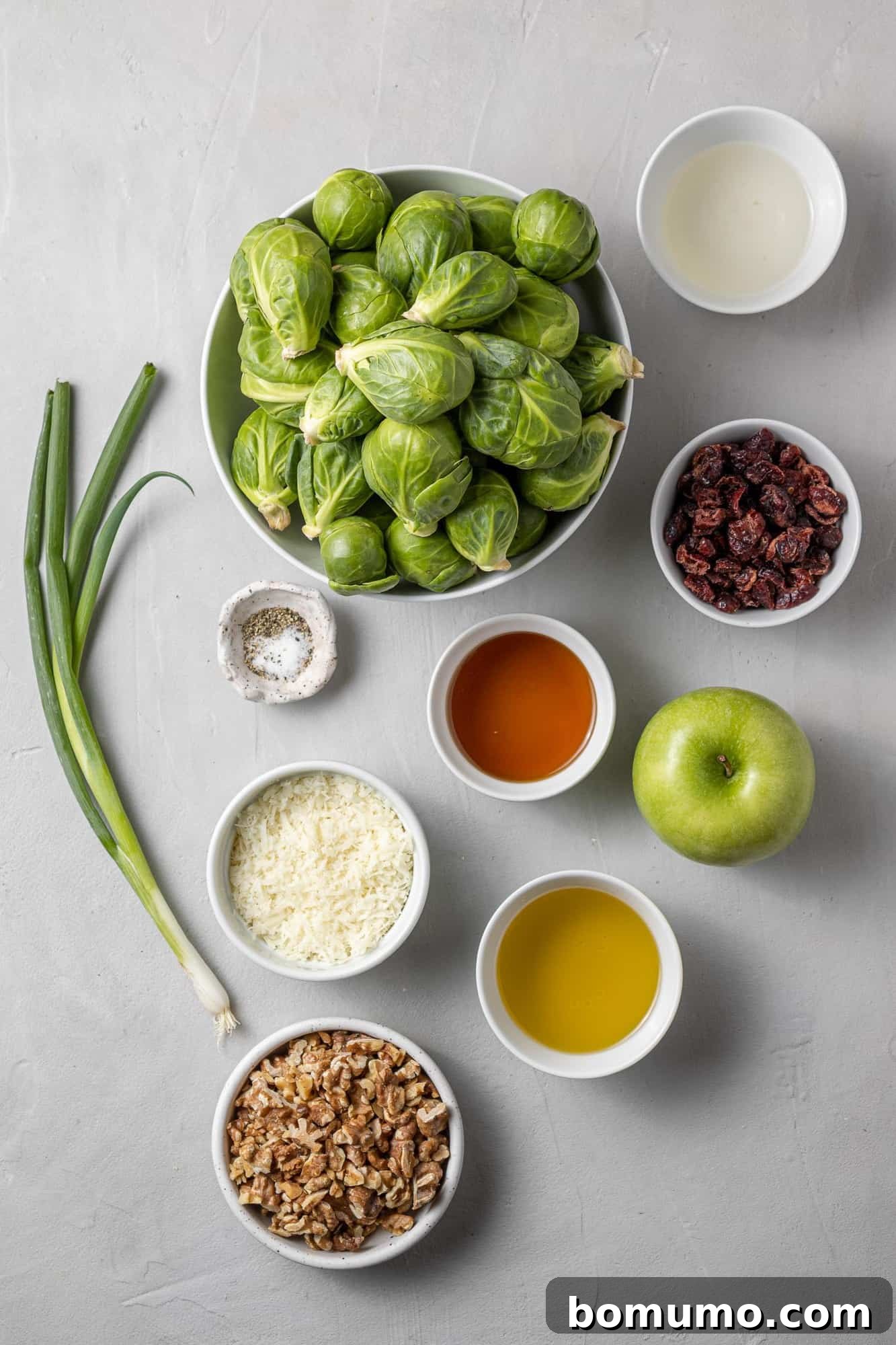 Overhead view of fresh ingredients including whole Brussels sprouts, green onions, walnuts, and a Granny Smith apple, laid out on a clean surface.