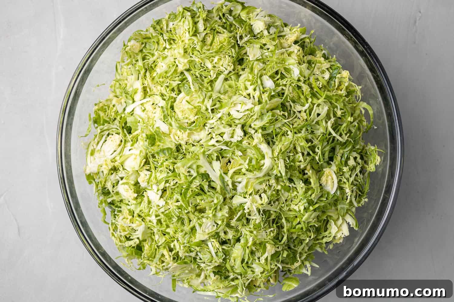 Shredded Brussels sprouts in a large clear glass mixing bowl, placed on a grey kitchen surface, ready for other ingredients.