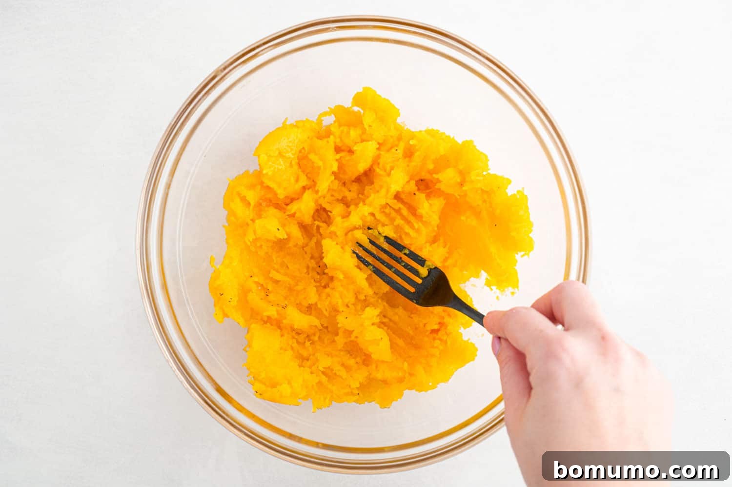 Squash flesh being mashed with a fork in clear bowl.
