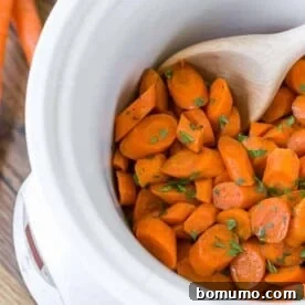Sweet and gently spiced crockpot glazed carrots in a white slow cooker, with a wooden spoon and a sprinkle of parsley.