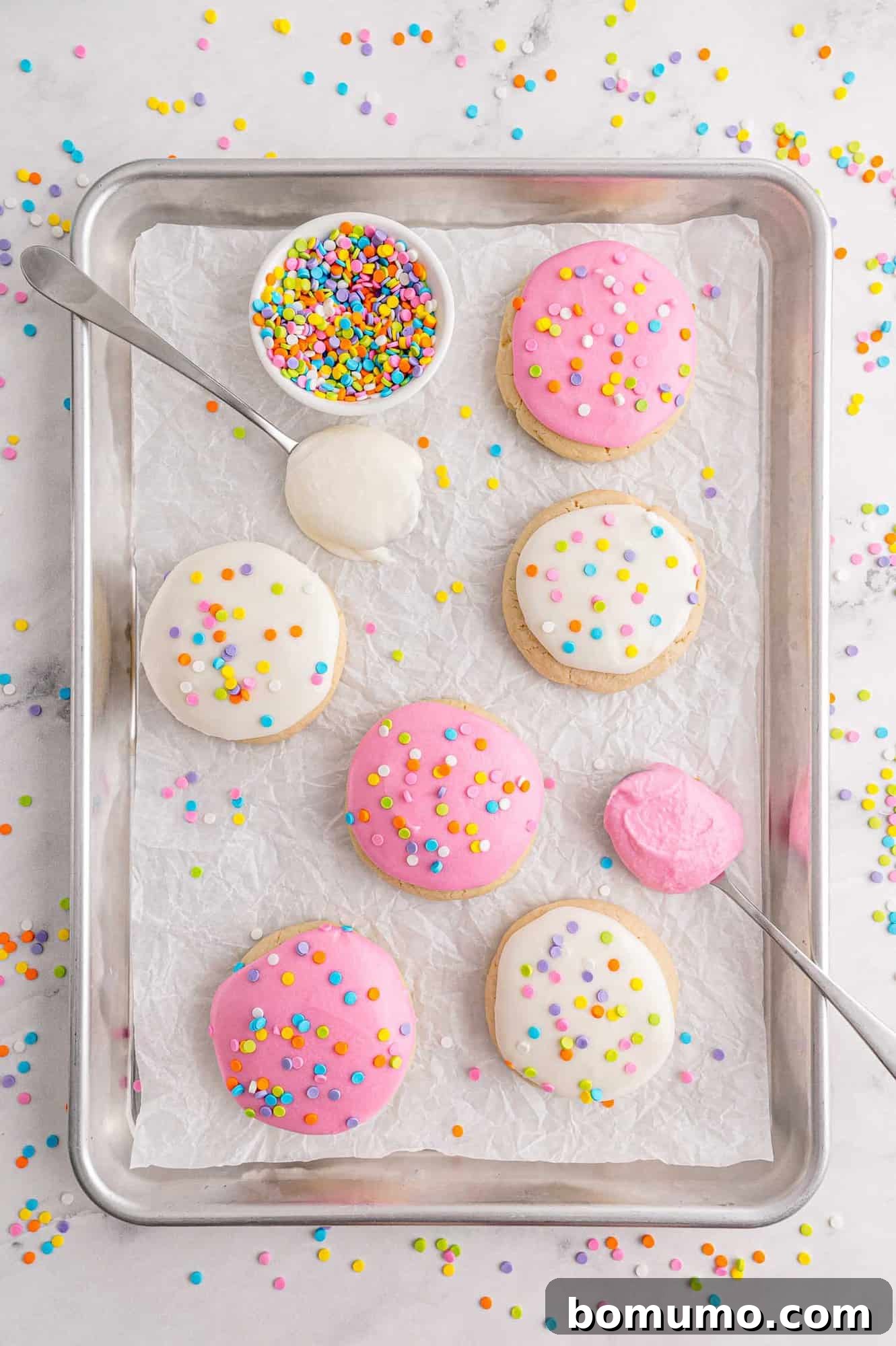 Tray of cookies with spoons full of frosting and a bowl of sprinkles.