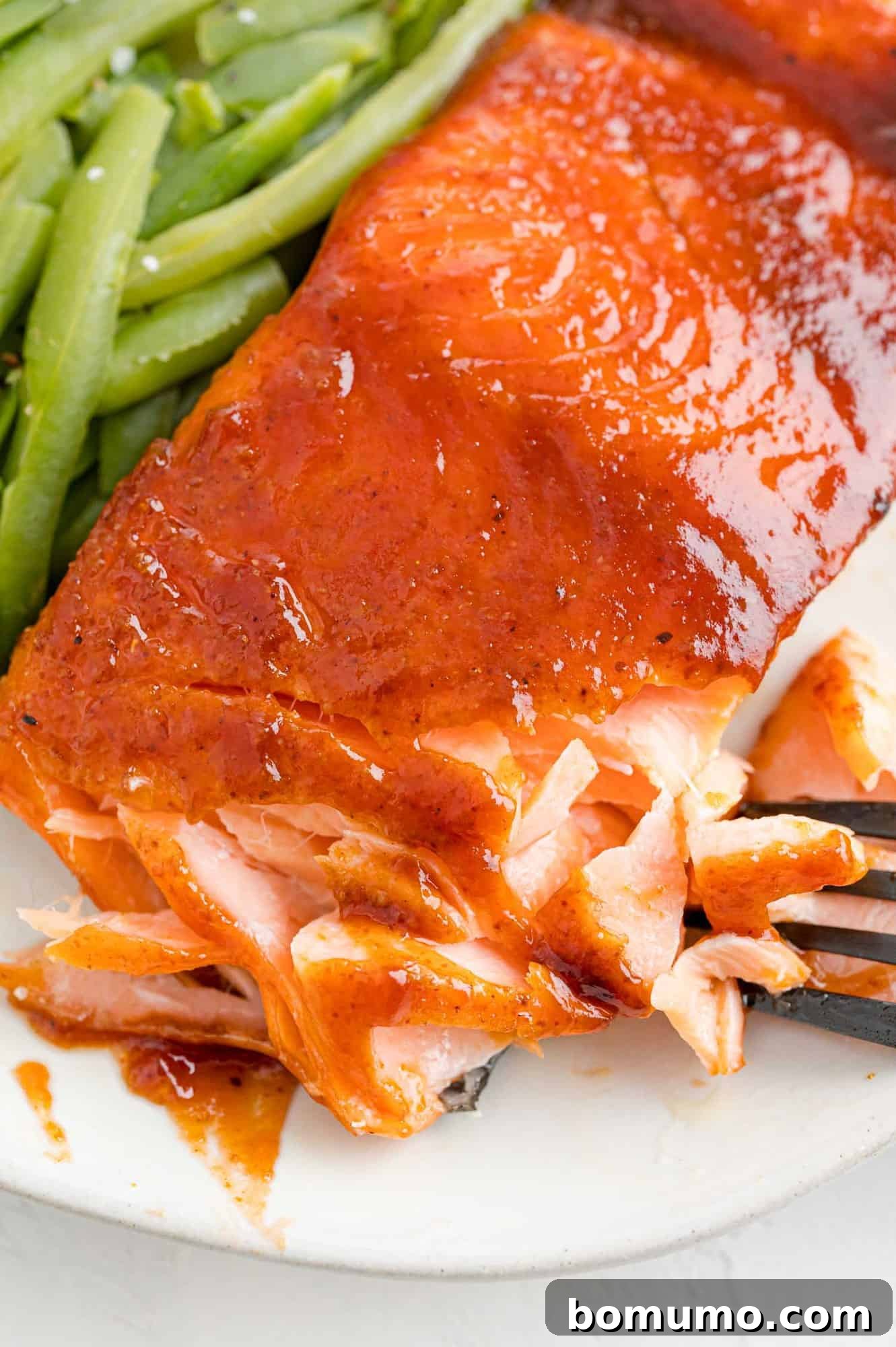 Savor the flavor of this quick and easy BBQ salmon, coated in a spicy dry rub and finished with a sweet, smoky barbecue glaze. Ideal for any weeknight, without the need for a grill! Perfectly cooked BBQ salmon next to vibrant green beans on a white plate, with a fork flaking the tender fish to show its juicy texture and rich color.
