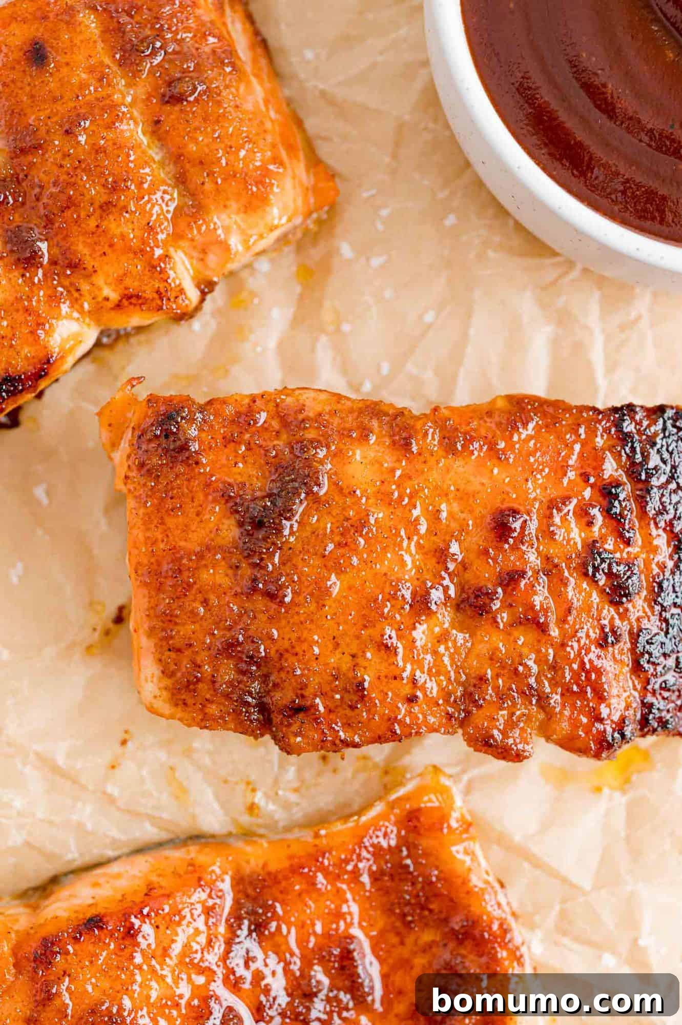 A feast of BBQ salmon fillets, beautifully prepared and ready for a savory dip in barbecue sauce. Three perfectly cooked BBQ salmon fillets on parchment paper, next to a small white bowl filled with glossy barbecue sauce.