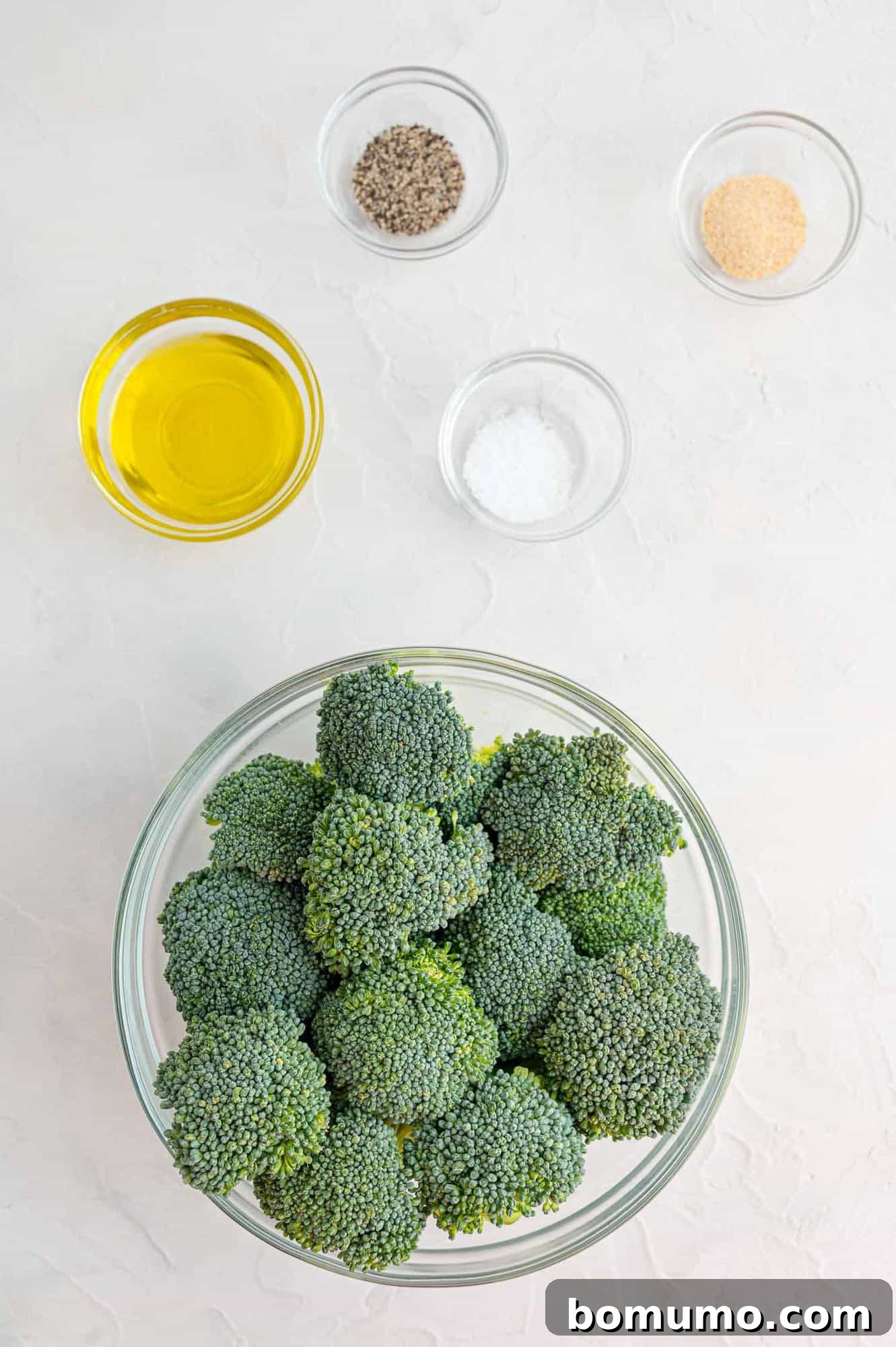 A selection of fresh ingredients laid out: vibrant green broccoli florets, a small bowl of golden olive oil, and white bowls containing salt, black pepper, and garlic powder.