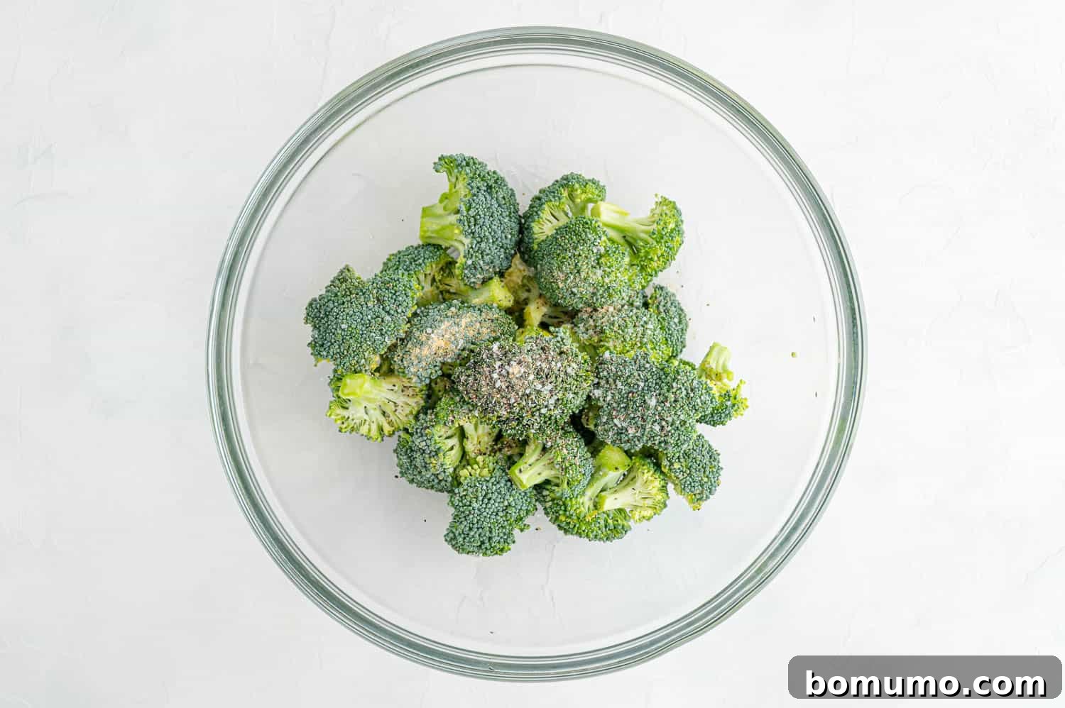 Broccoli florets in a clear mixing bowl, tossed with olive oil and a sprinkle of salt, pepper, and garlic powder.