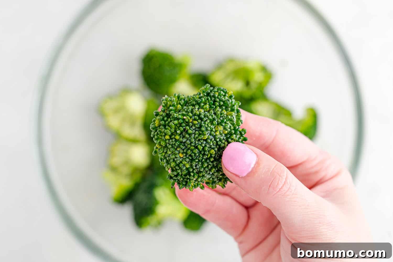 A hand gently tossing broccoli florets in a clear bowl, ensuring they are evenly coated with olive oil and seasonings.