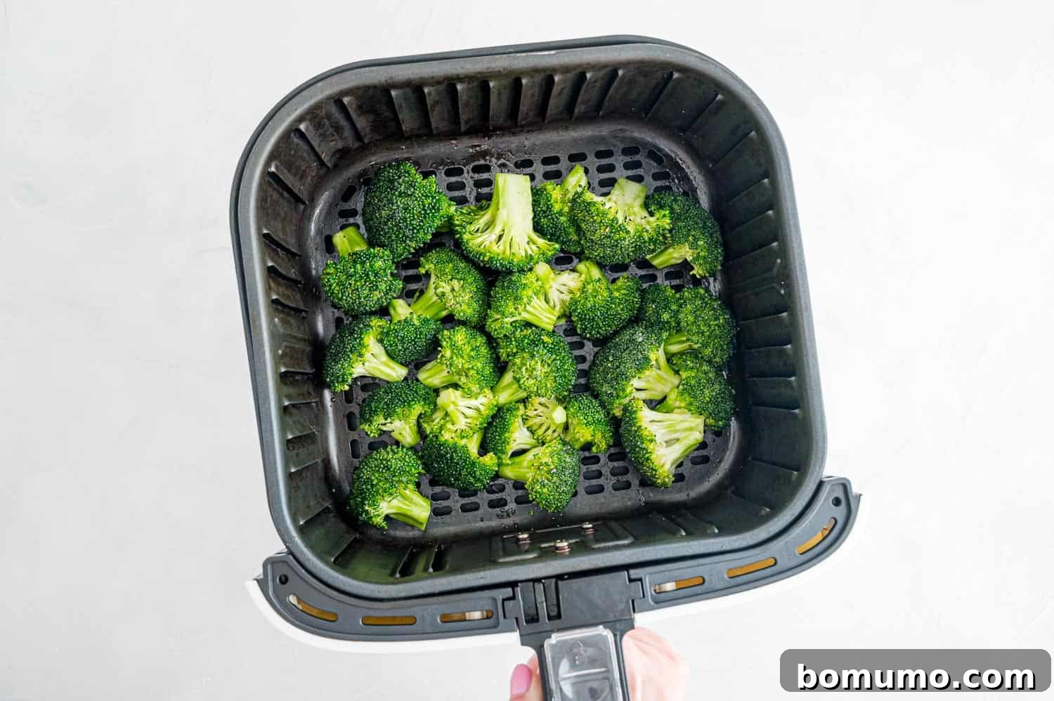 A hand holding and shaking an air fryer basket filled with seasoned broccoli florets, ensuring even cooking.