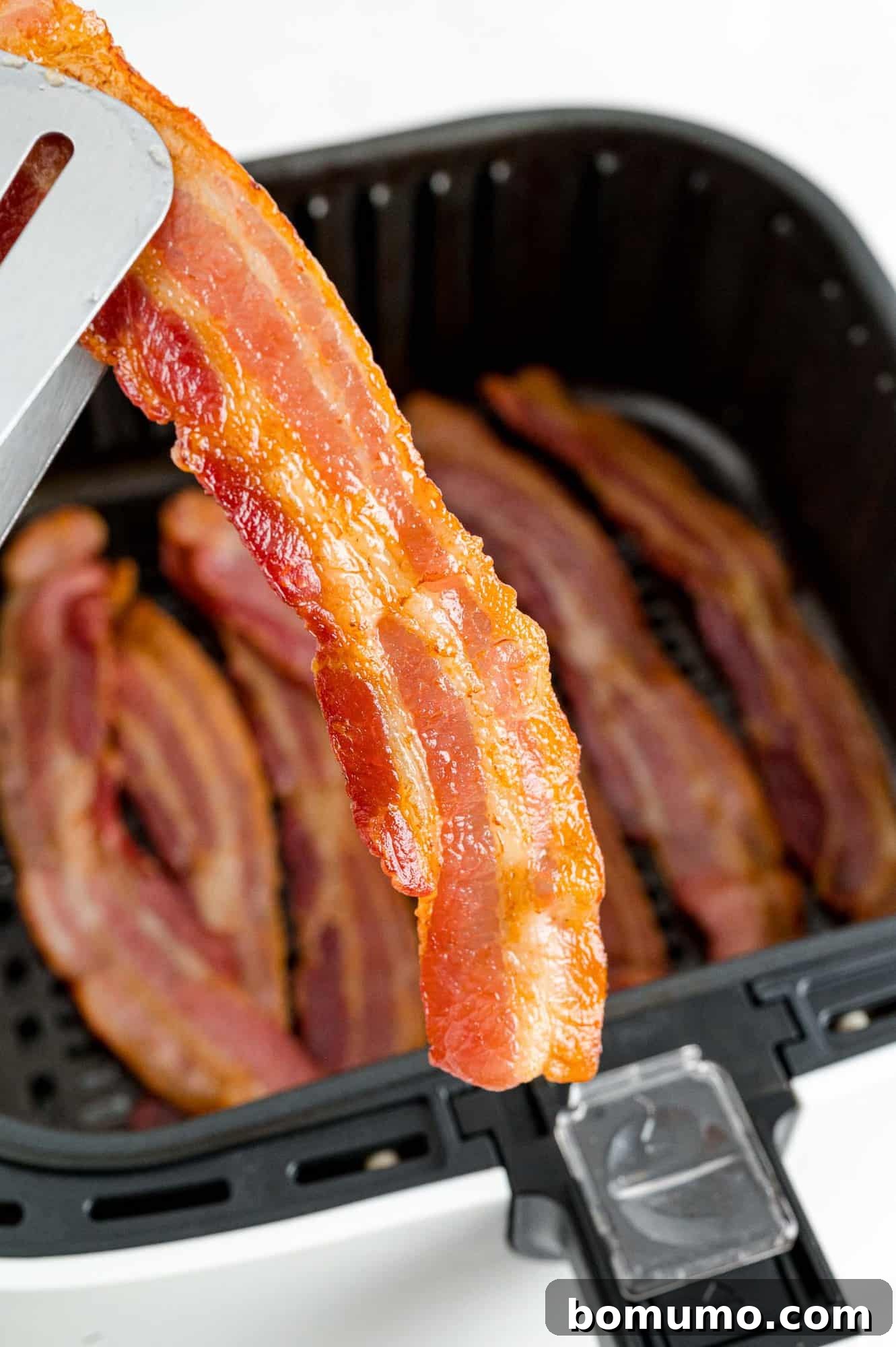 Crispy Air Fried Bacon 3 Cooked, crispy bacon being carefully removed from an air fryer basket with tongs.