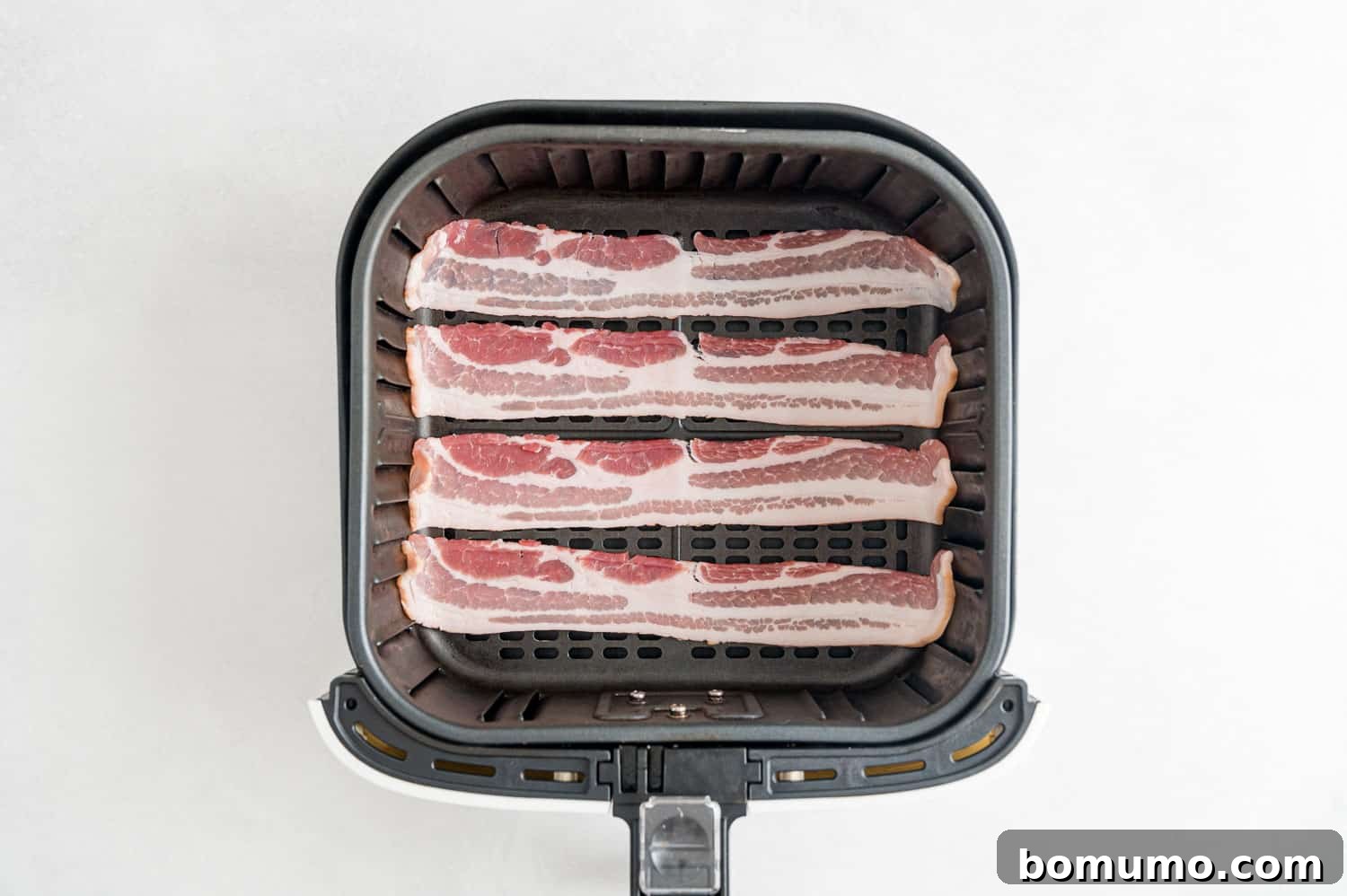 Crispy Air Fried Bacon 6 Uncooked bacon strips laid out in a single layer in an air fryer basket.