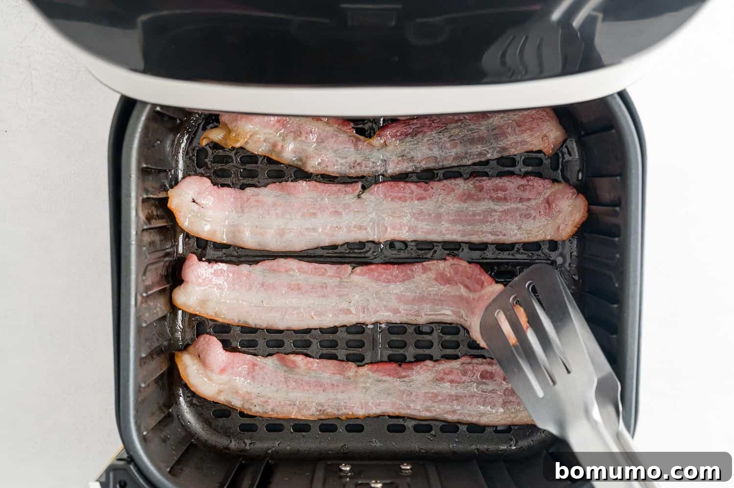 Crispy Air Fried Bacon 7 A close-up of bacon strips being flipped with tongs inside an air fryer basket.