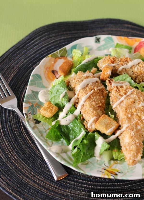 Ultimate Caesar Chicken Tenders 2 Crispy Caesar chicken tenders served on a bed of fresh romaine lettuce, drizzled with creamy Caesar dressing, on a decorative plate with a fork.