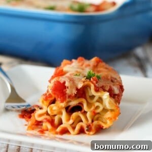 Lasagna pasta rolled up with pepperoni and cheese, coated in a tomato sauce. It is on a white plate and a blue baking dish appears in the background.