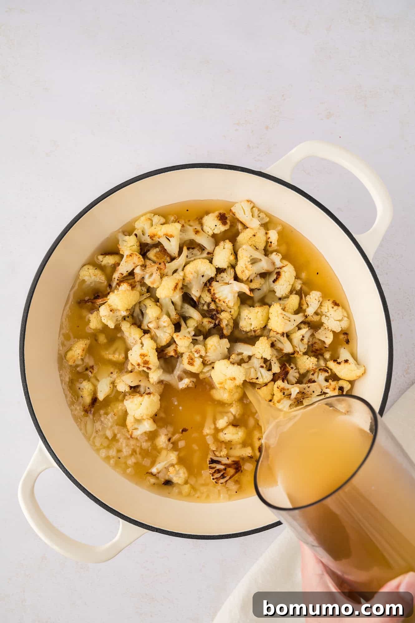 Rich, low-sodium broth being poured over the roasted cauliflower and aromatics in a Dutch oven, preparing the soup for simmering.