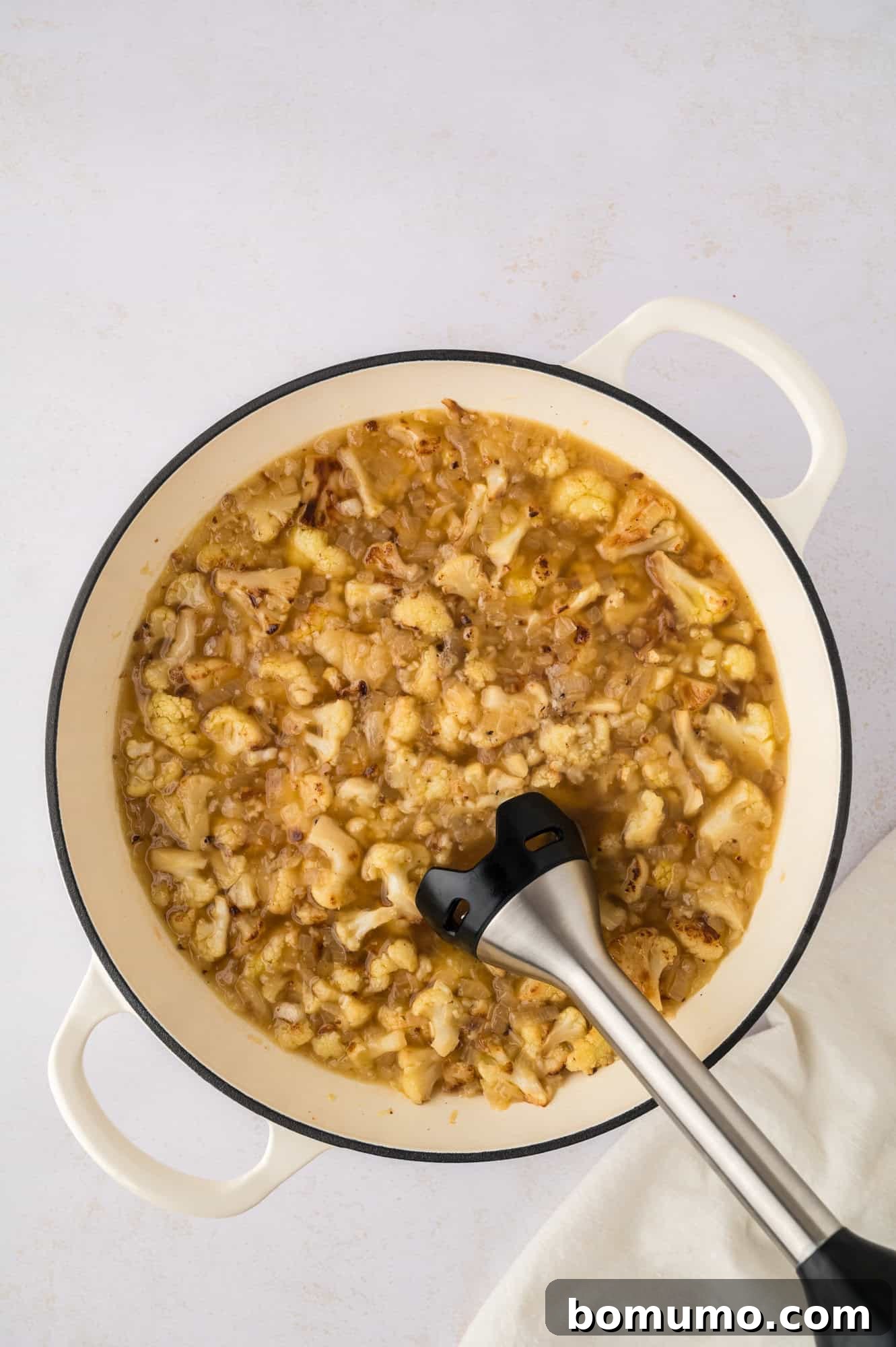 An immersion blender smoothly puréeing the roasted cauliflower soup directly in the saucepan, creating a creamy texture.