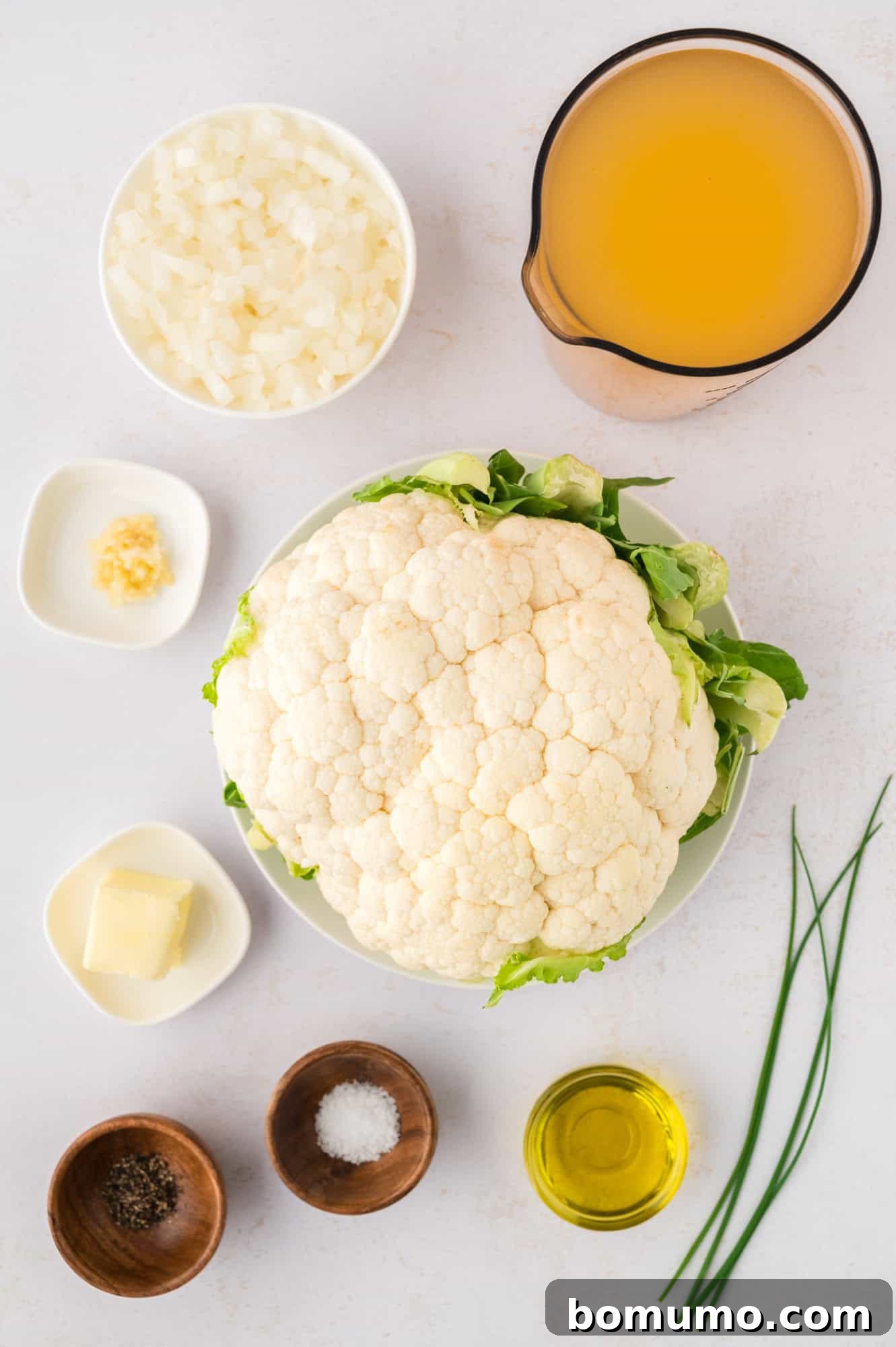 A vibrant display of fresh ingredients for roasted cauliflower soup, including a head of cauliflower, onions, garlic, broth, and butter.